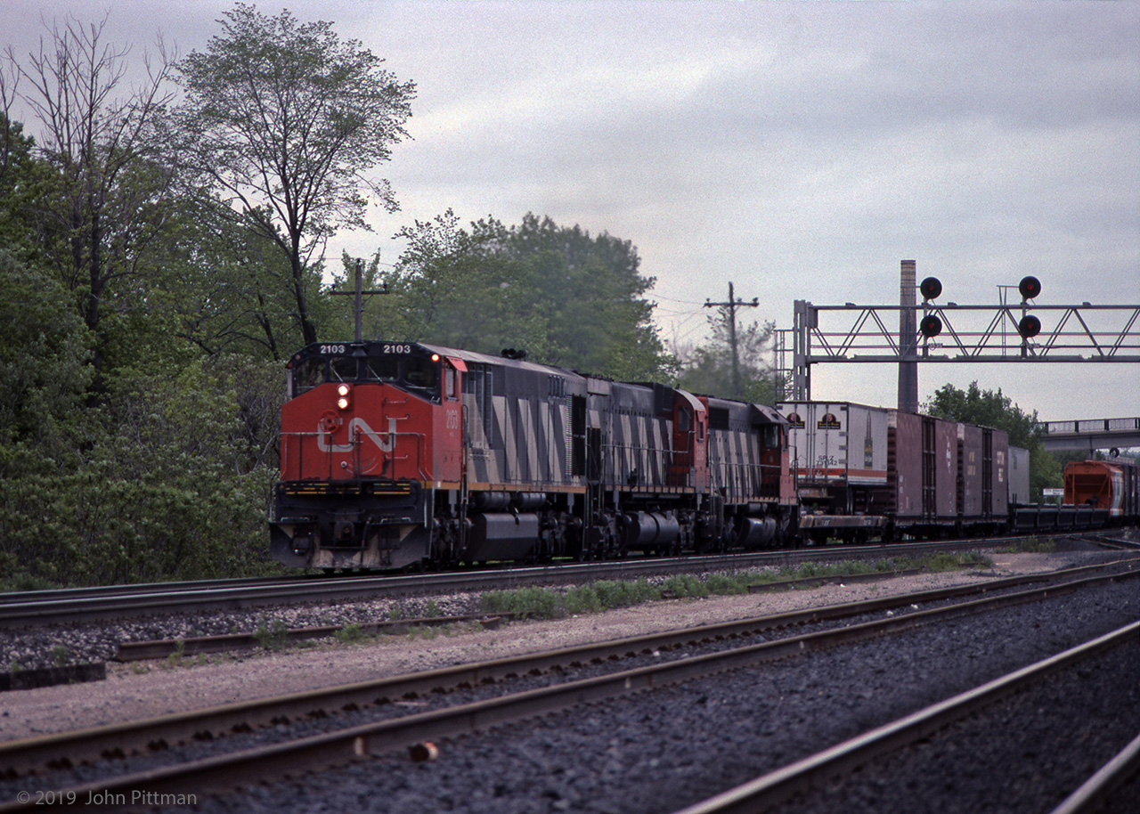A CN freight powered by HR616, M636, and SD40 is eastbound on the south track through CN Aldershot West, a year or so after the Aldershot Yard expansion moved its west-end connection here. 
Cars on the train include an orange and white Dupont Canada "sclair" covered hopper, 5 well-car platforms ahead of it, and a head-end flat car carrying highway trailer SPLZ 731342 with "Southern Pacific Golden Pig Service" - emblem is a smiling pig wearing an SP cap. 
Vantage point is the wrong side of the tracks from a public access point of view, times have changed. The chimney of the former Burlington Brick works, beyond Lemonville Road bridge, is further away than it looks.