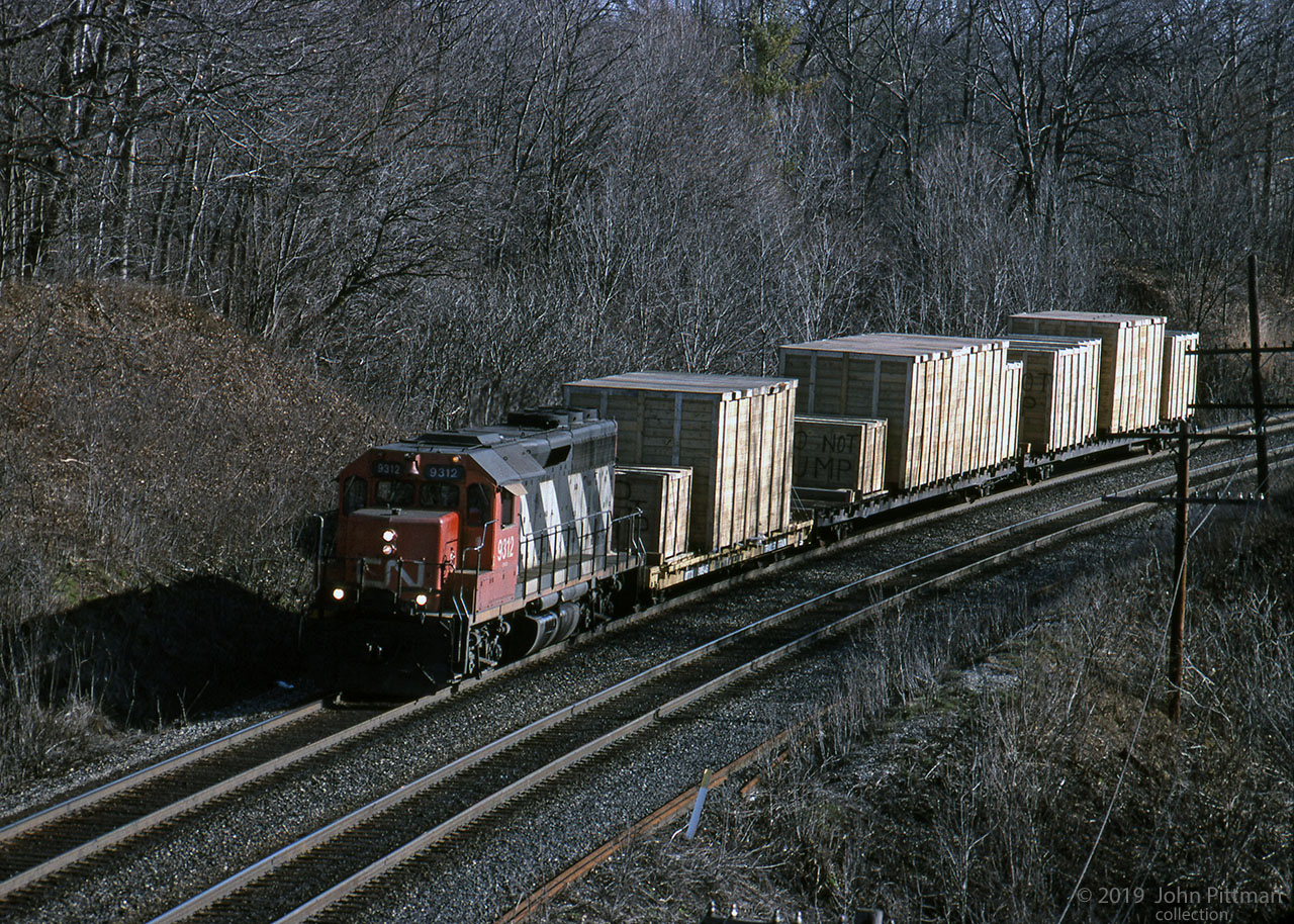 In early morning sunlight, standard cab GP40 locomotive CN 9312 leads a short train of 3 flatcars loaded with large wooden crates, contents unknown. 
The crates look large enough for this to be a "dimensional" train. "Do Not Hump" can be seen on some of the crates.
The train is westbound on the CN Oakville sub north track approaching CN Bayview (Junction); vantage point is sidewalk of Plains Road (Wolfe Island bridge).

Only 1 of the 2 "Extra" lights above 9312's number boards is illuminated, several other pictures by Bill also show just 1 of the 2 illuminated.
CN 9312 was built in 1967 by GMD London, original number CN 4012. The 4000-series GP40's were renumbered as 9300's in 1981, below the GP40-2(L)W range starting at 9400.