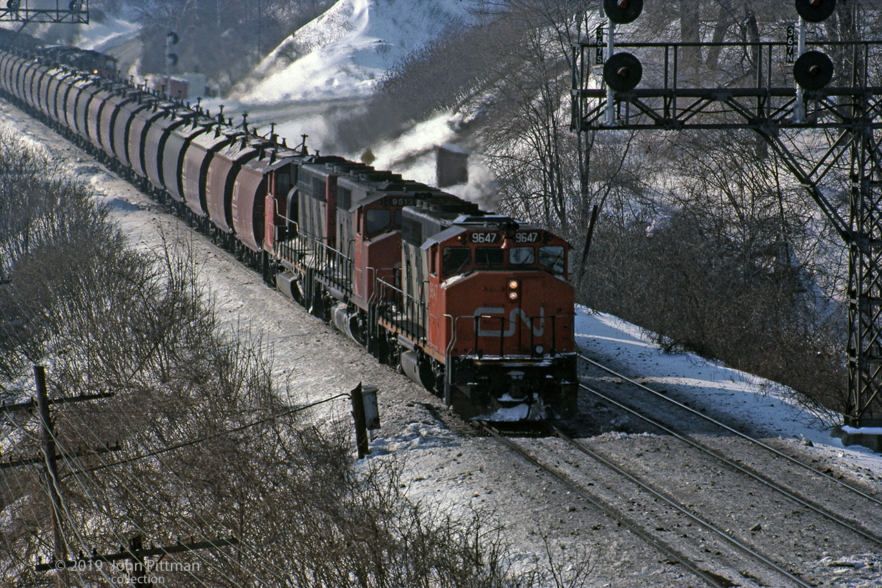 A trio of CN GP40-2W's haul a train of iron ore pellet cars uphill out of Hamilton, photographed on the Oakville sub south track between signals either side of CN Bayview. Presumably this eastbound train is empty cars heading back to load more pellets from the mine.
The upper part of another eastbound is visible on the north track near the far signal; the lead unit has a CN safety cab and is probably GMD, while the amount of dark smoke above the trailing units suggests MLW/ALco engines. 
The photographer wrote on the slide that this was a Dofasco train, and I've read that the iron ore came from  mines in Ontario's Temagami region, north of North Bay.