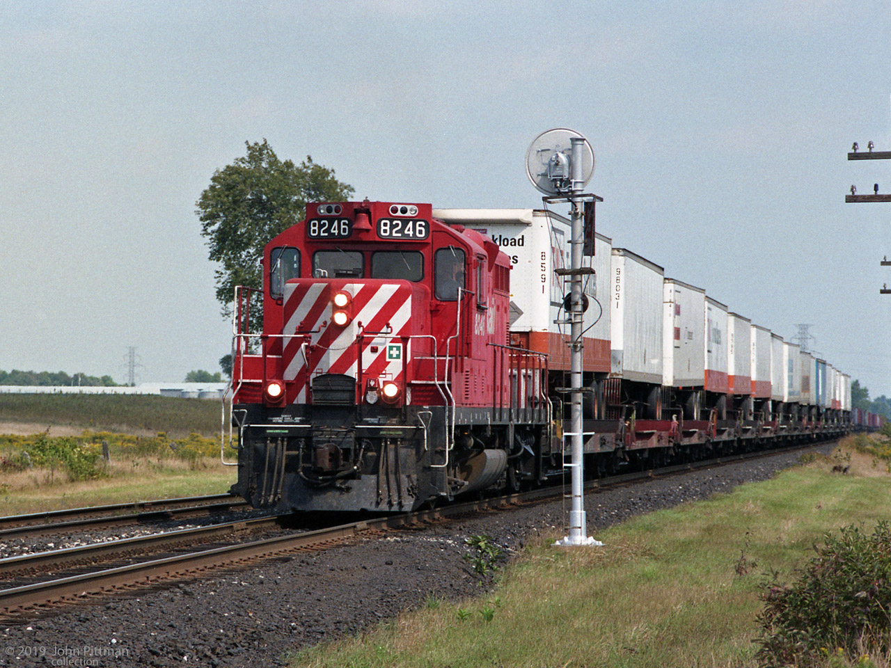 Remanufactured GP9u CP 8246 has a westbound train of at least 16 trailers on flat cars - the majority in TNT colours. 
It appears to be proceeding forward on the main after a signal stop that allowed an eastbound past on Nissouri siding. Part of the eastbound train can be seen at the right, beyond the piggyback train.
