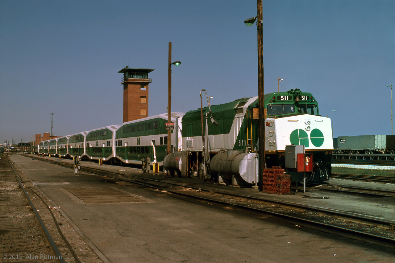GMD F40ph GOT 511 with its train of 7 multilevel 2000-series coaches and a single-level cab car sit out the weekend beside GO Transit's fueling facilities, in this east-facing view. The reddish brown objects stacked in front of the closest pole could be brake shoes.
Left over from the former CN Mimico Yard here on the north side are some buildings, with the east yard tower prominent. We signed our releases and borrowed hard hats in another. 
GO's F40ph locomotives were new in 1978, and GOT 511 is very clean and glossy. A CN person invited us into the cowl body engine room of an idling F40ph; he opened up a valve cover to show the exhaust valve rockers working and revved up the engine, a very loud and memorable experience.  
The main line tracks of CN's Oakville sub are on the white gravel at right. The red brick buildings beyond the end of the train are close to Mimico GO station. Don't expect to be permitted into any rail yard nowadays - however Islington Ave bridge provides good views of the GO and VIA yards.