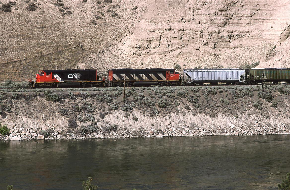 A westbound CN grain train passes by an interesting slope. Grazing animals created the converging line on the hill at left, while rivers and lakes created the interesting bedding in the poorly consolidated "rock" formation at right.