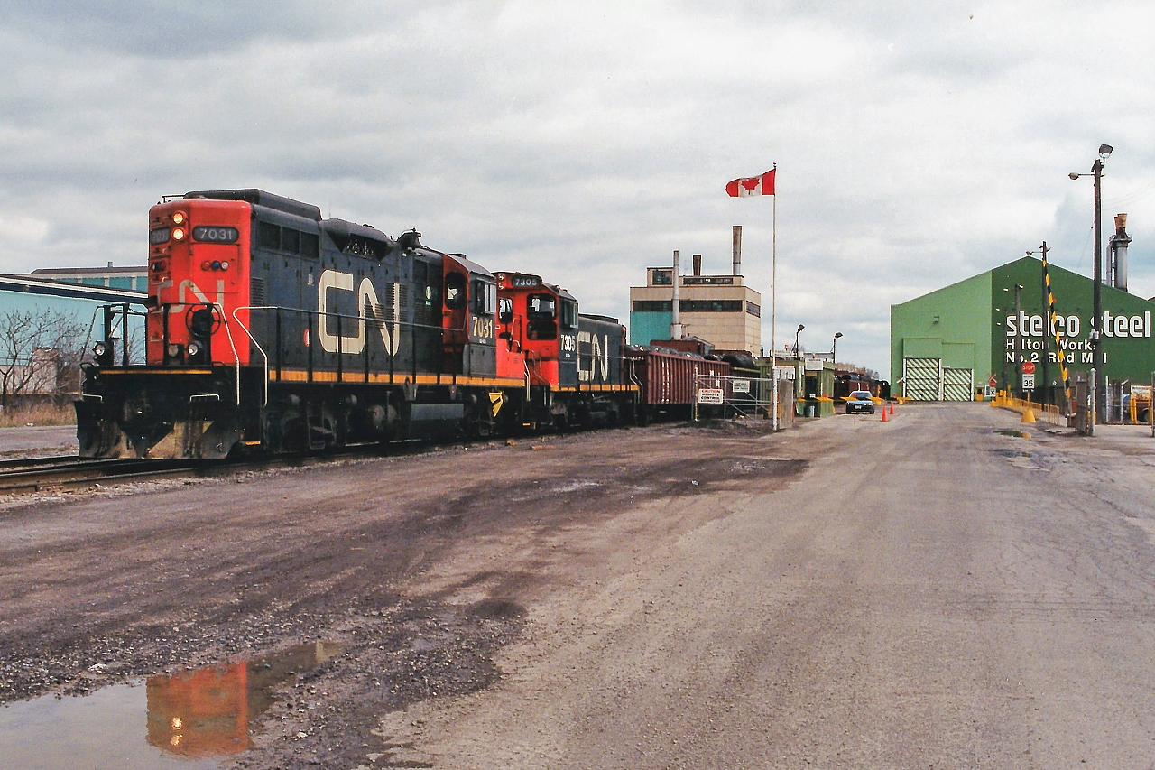 Taking a break from mainline trains, two friends and I ventured down into Hamilton’s Ontario industrial core for the first time during a spring morning in 1995. I remember at the time we were overwhelmed with the maze of tracks and all the industries that were still served by rail as well as all the industrial units that were working behind the seemingly endless rows of fences. However, not long into our tour, we came across CN GP9RM 7031 and SW1200RSM 7305 switching the Stelco Steel Rod Mill facility. We took a few photos and also shot the pair working the nearby Stelwire Parkdale plant. I believe that at least one of these industries still receives rail service today; however corrections and additional information are both welcome and would be appreciated.