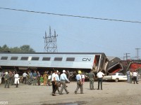 A crowd of bystanders take in the cleanup efforts of the wreckage of CN Tempo train #150, derailed along the Oakville Sub one mile east of Oakville station while travelling from Sarnia to Toronto. A wreck train and big hook work to clean up Tempo cars that are sprawled along the mainline, some coming to rest on automobiles parked in this adjacent lot. RS18 3151 is pictured on its side in the distance. One woman was killed while walking between cars when the train derailed, eight people were injured, and the engineer lost an arm.