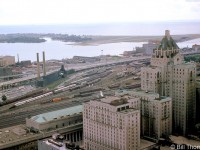 Here's an aerial view of the downtown Toronto railway lands taken from the Bank of Commerce building's observation deck in September 1964. Visible in the foreground is Toronto's Union Station on Front Street, opposite the CPR's famed Royal York Hotel.<br><br>Behind the station's train sheds are CPR's John Street coachyards, roundhouse and servicing facilities for both locomotives and passenger cars (CN's Spadina facilities are out of frame to the right). A switcher is visible moving around stainless steel Budd equipment for The Canadian, amid plenty of heavyweight and lightweight steel coaches in the yard. A pair of F-units can be seen on the servicing tracks near the roundhouse by the old coal tower, along with a pair of Budd RDC cars ("Dayliners" in CP parlance). Most of the view of the roundhouse is blocked by the top of the Royal York.<br><br>Nearby is the Toronto Terminals Railway Central Heating Plant (which supplied steam heating to downtown buildings including Union Station, the Royal York, and CP's John Street facilities). Along the harbourfront are buildings for Yardley's (perfumes), the Terminal Warehouse, grain elevators, and various piers. The Toronto Island airport is off in the upper right.