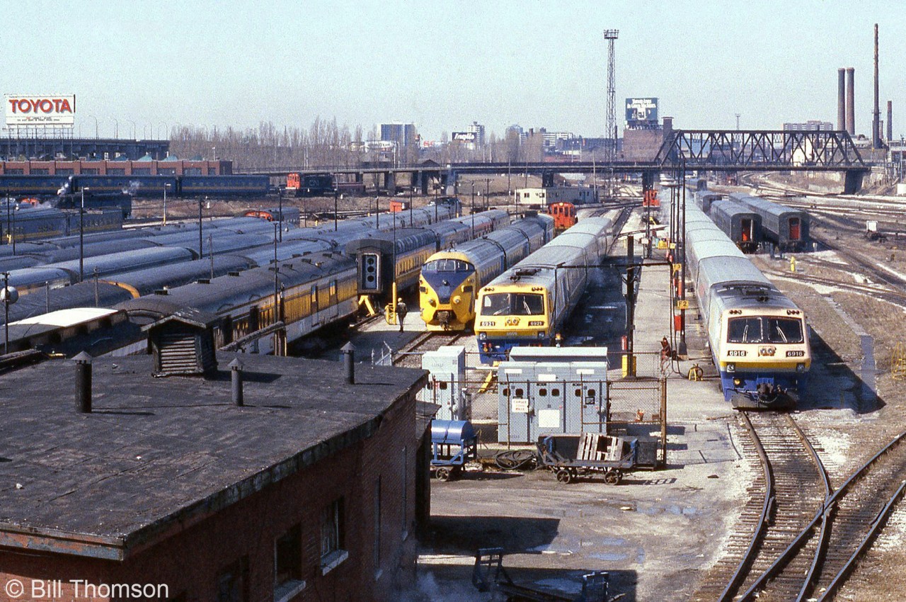 VIA's (formerly CN) Spadina Coachyard in Downtown Toronto is pictured in March of 1984. The tracks are teaming with passenger equipment, including VIA LRC consists with locomotives 6916 and 6921, an Ontario Northland TEE train for the Northlander, Tempo cars, cars from the blue and yellow fleet, a steam generator car, and old heavyweights including some baggage cars still painted in CN colours. All of this was switched by a fleet of CN S13 units, five of which are visible. They also shuttled entire consists to and from nearby Union Station.

By this time in the mid-80's all of the former CN steam-era facilities and yards at Spadina were on borrowed time. Soon VIA would move operations to nearby Mimico (the new Toronto Maintenance Centre and yards), and the downtown railway lands would be cleared in preparation for construction of the new Skydome (Rogers Centre) where the old Spadina roundhouse was. Most of the former coachyard property would lay empty until development years later, into mostly condos.