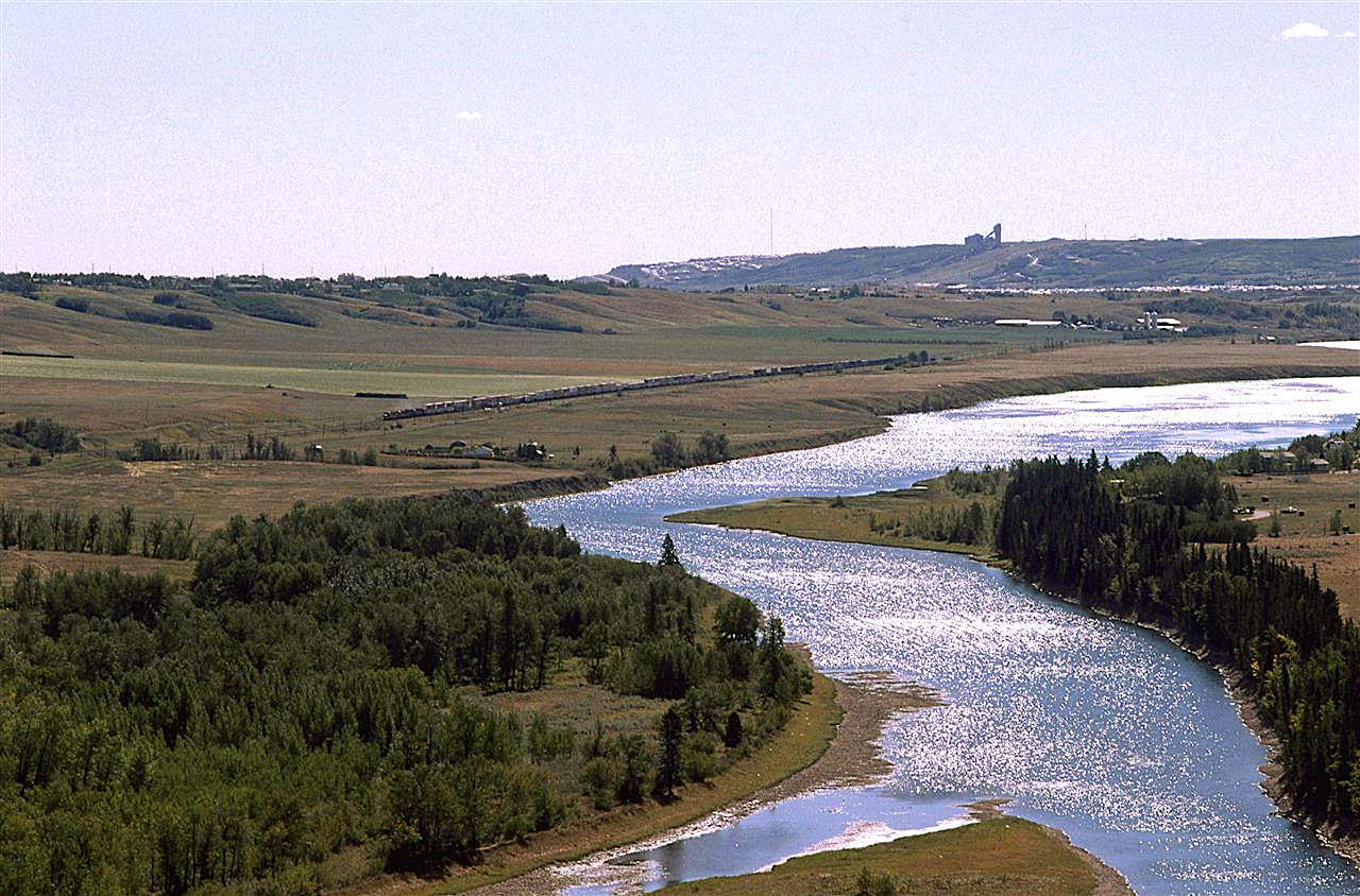 A westboind intermodal train comes out of Calgary along the Bow River at the westernmost end of Bearpaw Reservoir. Calgary Olympic Park facilities and urban sprawl can be seen on the horizon
