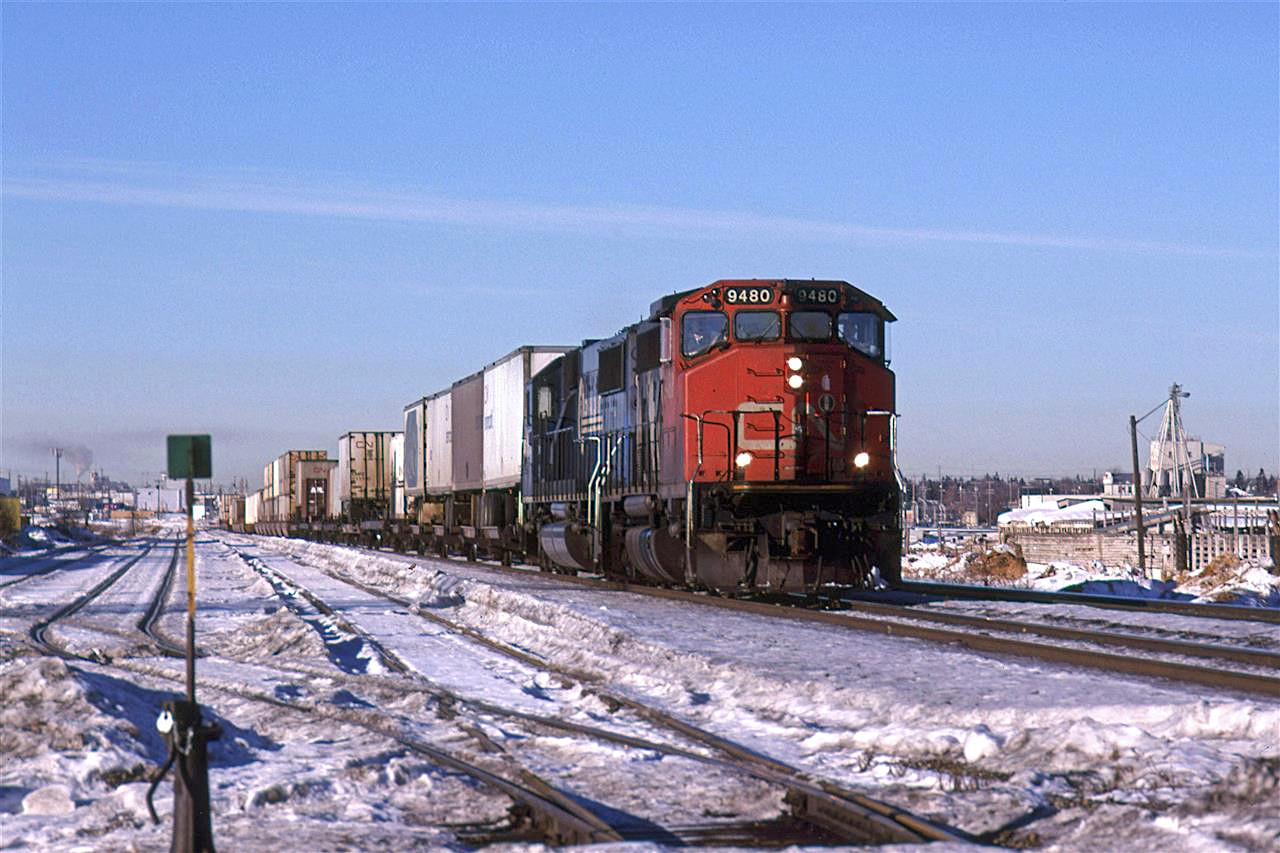 It must be the February thaw in Edmonton.
Besides the train with the Conrail unit trailing, there is quite a bit to see here. At far right, the stockyards can be seen. A sma;; specialized elevator is al so visible. At right, the smoke/steam is from the inland cement plant on the far side of town