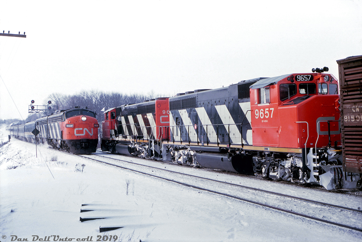 It's a little nippy out on this cold morning at Bayview Junction, as less-than-a-year-old CN GP40-2W units 9658 and 9657 in charge of train #409 meet eastbound VIA Rail passenger train #70 with FP9 6528 and FPB4 6866 in the lead.You might not know it, but this photo was taken just days after VIA Rail as it currently exists was formed: CN's VIA branding had started in 1976, but VIA officially became a Crown corporation just three days earlier on January 12th, when CN (still a Crown corporation itself) spun off its passenger operations into a new separate entity. Here, the power and most of the train still sport CN colours, but a VIA-painted coach is visible on the head end. Over the next few years, most of the former CN & CP passenger equipment VIA acquired would be repainted into the common blue and yellow scheme (with a few exceptions, such as old equipment retired early on, and a few stragglers like an FPB4 that lasted in zebra stripes until retirement).Checking a 1976 timetable, if my calculations are correct train #70 would have hit Bayview Junction around 9:15-9:25am in the morning, if running on schedule. Being inside riding the steam-heated passenger cars was probably quite nice on days like this!Reg Button photo, Dan Dell'Unto collection.