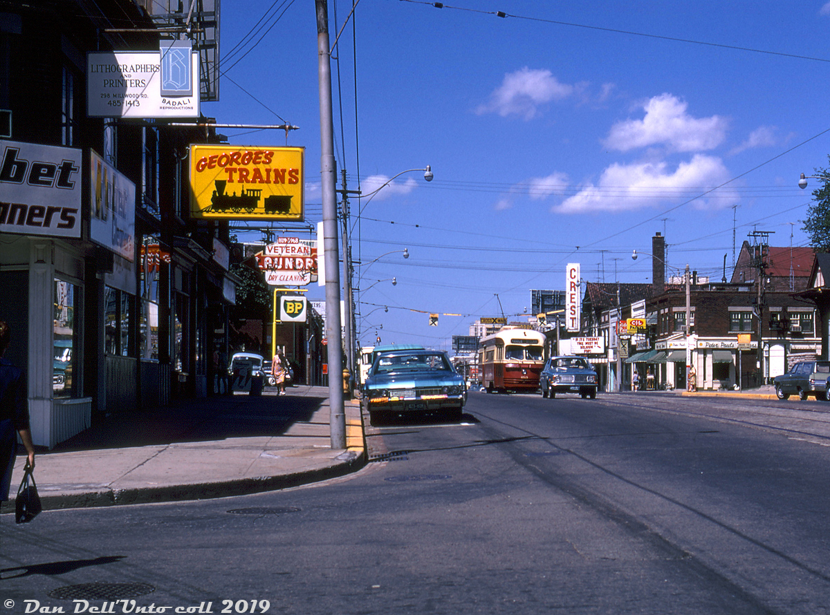 A late afternoon in the Davisville/Mount Pleasant area of Toronto finds TTC PCC 4515 in traffic heading southbound on Mount Pleasant approaching Millwood, operating on the St. Clair route heading "westbound" from Eglinton to Keele Loop. This would be a rather unremarkable shot, except for the inclusion of the famed George's Trains store, back when you could literally ride the rails to their doorstop.

In the 1940's and 1950's this location was known as Bob's Hobby Shop (owned by Bob Goodall, and later Bob's Toy Trains), at 510 Mount Pleasant Avenue. The story goes that George Olieux (the founder of George's Trains) did model repair work for a few shops in the area, including Bob's. In the early 1960's he bought out Bob's store, and the rest was history. For decades, many generations of model railroaders, railfans, and transit enthusiasts made the trek out to George's to get their model train fix (or magazine and trackside guide fix), either by driving, transit or even on foot (Millwood is a nice walk in the fall from the nearby subway). Streetcar service on Mount Pleasant was discontinued in June 1976, replaced by trolleybuses, which in turn were replaced by the present diesel bus service.

George passed away in 1995, and the business was continued by his sons. In 2007, George's Trains acquired a second location in Markham (the former Railview Trains store, owned by the late David Morgan). Alas, the old Mount Pleasant location was closed in May 2009, apparently due to a number of issues many businesses operating in Toronto have been facing in recent years (including higher taxes, increased traffic congestion, and lack of suitable parking). Operations continue at their suburban Markham location.

Photographer unknown, Dan Dell'Unto collection.

Opposite side along Mount Pleasant at Millwood: http://www.railpictures.ca/?attachment_id=32380