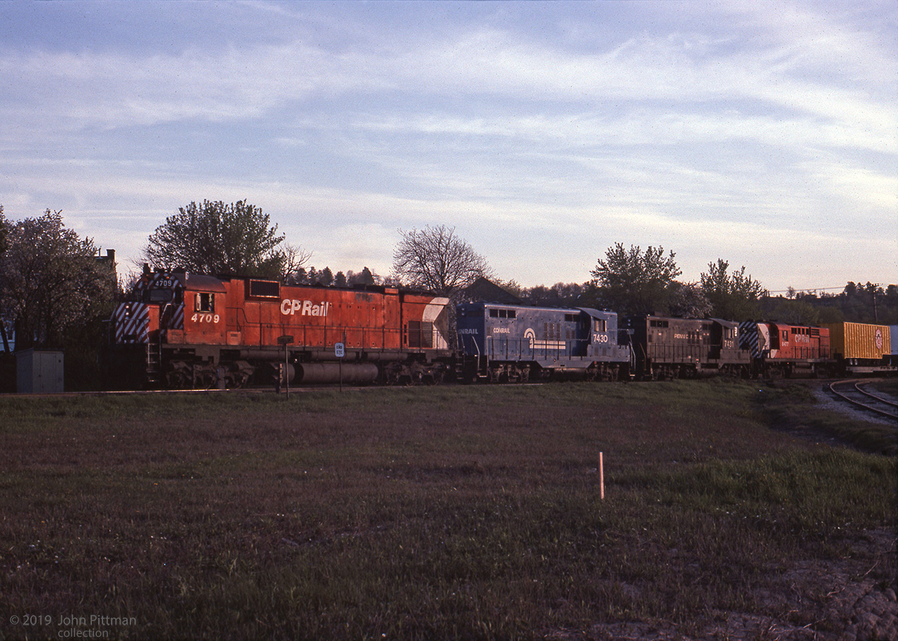 Big MLW-ALCo CP 4709 leads a pair of Ontario-based Conrail (PC) GP9's, RS10 CP 8477, and train close to sunset.  The first piggyback trailer has the Conklin Shows clown face logo (CNE midway, etc).
Clues to the location are signs "Yard Limits" and "End CTC", a dwarf signal, and the elevated right-side background.
Some experienced Hamilton railfans told me it looks like the "Starlight" train, facing toward Toronto, at the (north) east yard limit of Aberdeen. In which case, the diverging track at right could lead to the Chatham Street roundhouse area.