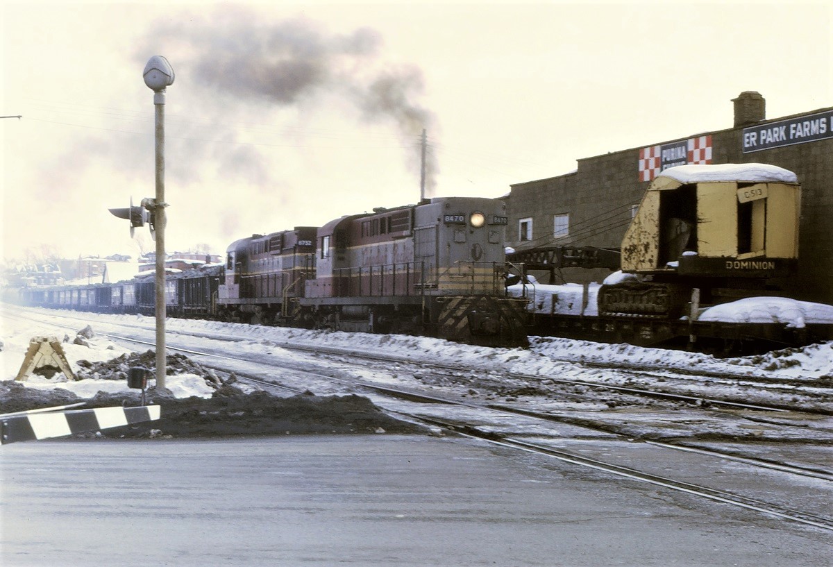 On a very overcast February afternoon, CP RS-10 8470 and RS-18 8732 have their work cut out for them as they pull an over tonnage train west on the Cartier Sub in downtown Sudbury, Ontario.  Out of sight is S-2 7090 shoving hard on the caboose to help keep the train moving.  The crane on the flat car beside the locomotives looks like it has had a pretty rough life.