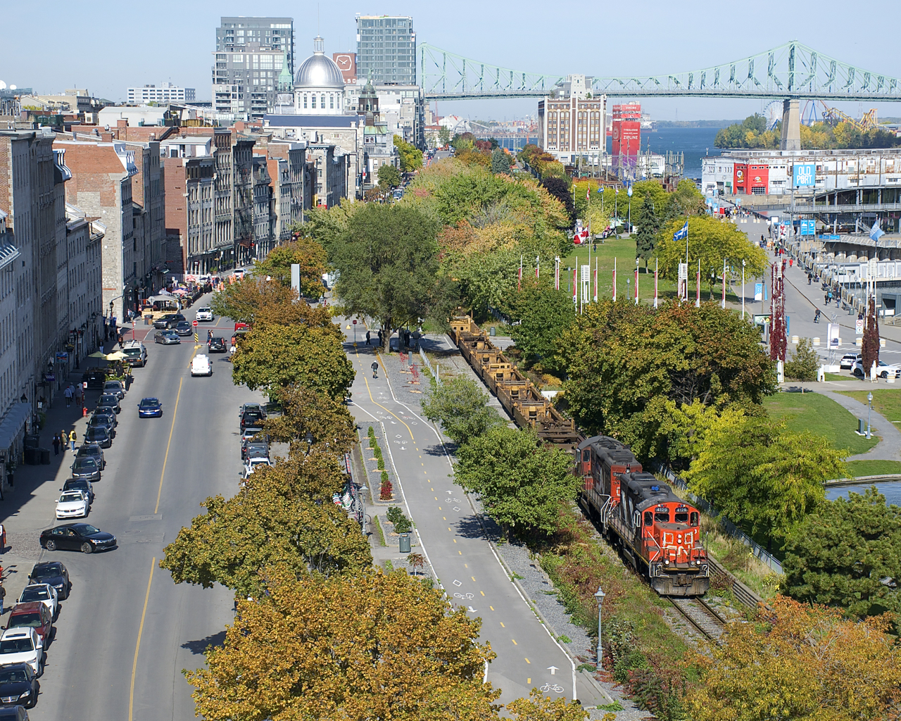 A CN transfer powered by CN 4129 & CN 7229 is exiting the Port of Montreal, with well cars up front and mixed freight out of sight further back. Paralleling the CN Wharf Spur here is a bike path and De la Commune Street.