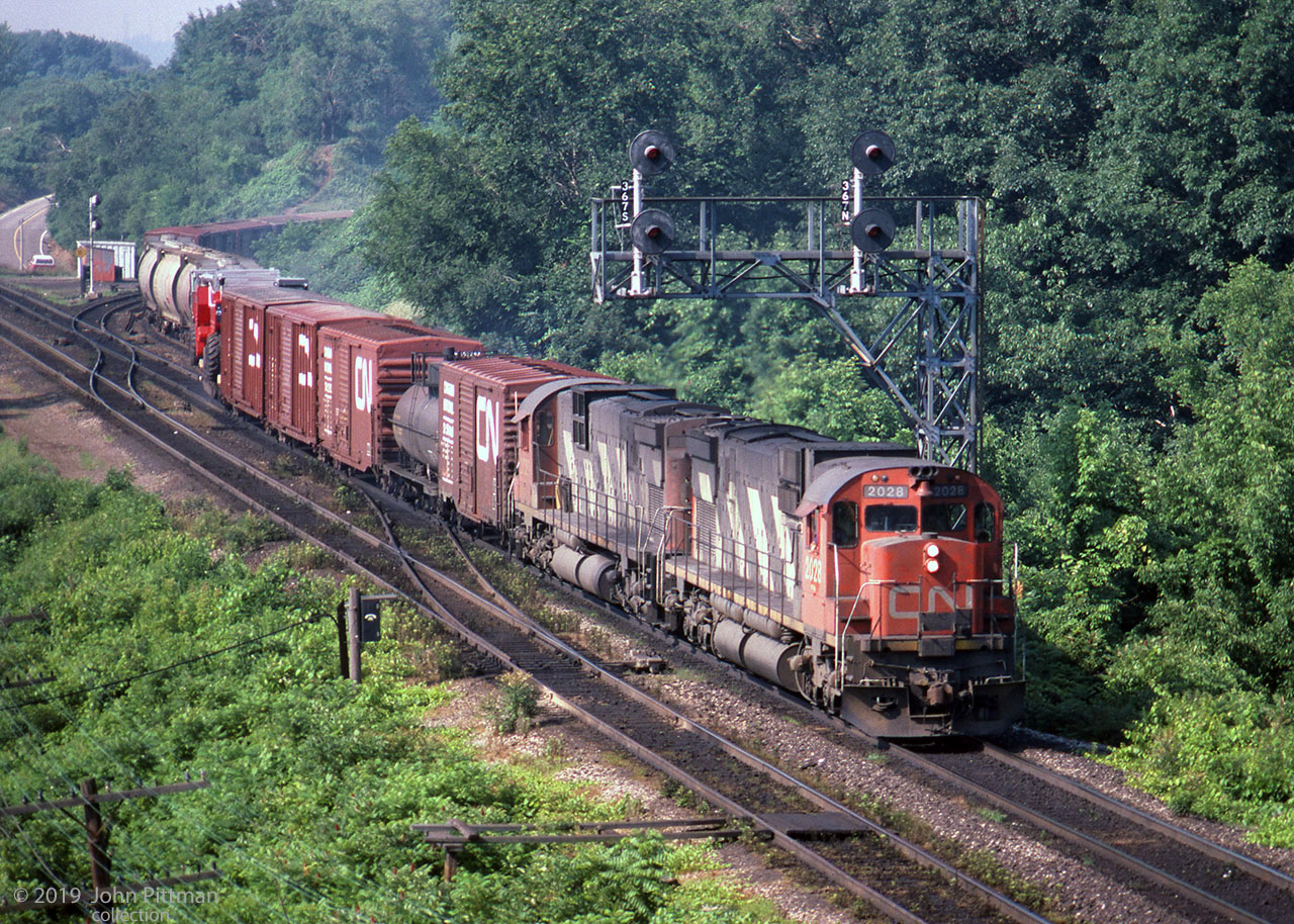 A morning CN eastbound powered by a pair of MLW C630m locos enters CN's Oakville Sub off the Dundas at CN Bayview.
CN 2028 and CN 2029 lead the train - the flatcar load 6 cars back looks agricultural, could be a combine harvester.
Of interest beside the south track are active lineside wires and poles, a CN telephone, and a pair of light rails perpendicular to the track - presumably to park a speeder.
The Hamilton/Burlington boundary is approximately where the engines are.