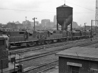 CN Locomotives spotted between Spadina Avenue and the Spadina Roundhouse in this mid-1962 picture.<br>
The visible engines still have CNR maple leaf paint schemes that preceded CN black and orange-red.<br>
Beyond MLW RS10 units CN 3107 and CN 3109 are MLW S-4 CN 8145 and GMDD SW1200 CN 7025. <br> 
Similar switcher exhaust stacks can be seen in the row behind the switchers, then the cab of an MLW RS-3.<br>
Near left is the long hood front end of a Grand Truck Western EMD GP9.<br><br>
With steam engines out of service, the ash pits are covered (between rails across from 3107's cab) and the ash conveyor structure is gone.<br> 
Partly obstructed by the left side of CN's water tower is the 34 story Bank of Commerce Building, for many years the tallest building in Toronto, in Canada, and in the British Commonwealth.