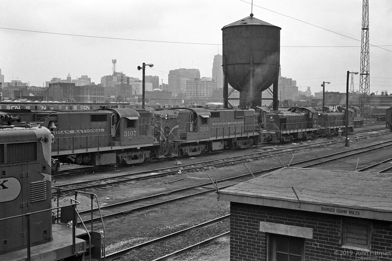 CN Locomotives spotted between Spadina Avenue and the Spadina Roundhouse in this mid-1962 picture.
The visible engines still have CNR maple leaf paint schemes that preceded CN black and orange-red.
Beyond MLW RS10 units CN 3107 and CN 3109 are MLW S-4 CN 8145 and GMDD SW1200 CN 7025.  
Similar switcher exhaust stacks can be seen in the row behind the switchers, then the cab of an MLW RS-3.
Near left is the long hood front end of a Grand Truck Western EMD GP9.
With steam engines out of service, the ash pits are covered (between rails across from 3107's cab) and the ash conveyor structure is gone. 
Partly obstructed by the left side of CN's water tower is the 34 story Bank of Commerce Building, for many years the tallest building in Toronto, in Canada, and in the British Commonwealth.