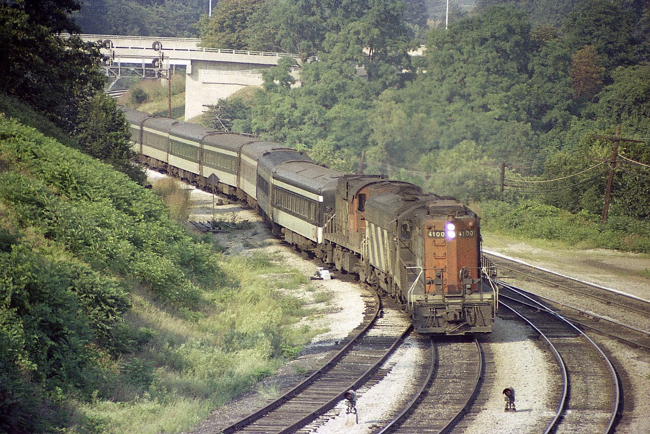This view from the hillside at Bayview Junction features CN 4100, 6630 and 3100 about to cross from the Oakville Sub to the Dundas and begin the long climb westward. It was a hot and steamy late August day, back before the formation of VIA, and anything could be seen powering the passenger trains.
Note the switch is still in for that "helper track" on the left, its purpose being that helper engines were stationed there to help heavy trains climb the grade to Copetown. The 'railfans walkbridge' in the background is brand new, but no buffs there; the heat probably drove them to cooler locations.......or home.