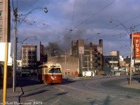 <b><i>Dundas Square, 1970's style</i></b>. TTC PCC 4384 operates on an eastbound run of the Dundas route, shown at Dundas & Victoria amid some morning unrest: smoke from an unspecified fire downtown (not the famed Eatons fire that took place a number of years later) is visible in the background, and whatever the incident was probably necessitated diverting Dundas cars down to Queen and across to get around it. 4384 is shown on the short connecting track between Victoria and Dundas proper, meaning it has just come up from Queen via Victoria, and is resuming regular routing on Dundas bound for Broadview Subway Station. Due to the lack of foot and auto traffic, it's possible that this was taken on a weekend.
<br><br>
Everything in this scene has changed dramatically in the years and decades that followed, and anyone frequening this area of Toronto today would be hard-pressed to find much of old still recognizable (the big <a href=http://www.railpictures.ca/?attachment_id=16283><b>10 Dundas Street East blob</b></a> doesn't help). Behind the streetcar to the right are the original Dundas Square retail/commercial buildings that once occupied the site, bounded by Yonge, Dundas, Victoria and Dundas Square. Tenants included Cole's (books), a Howard Johnson's, and the Downtown Theatre (whose street-side marquee signage is visible poking up). The whole "square" of buildings was demolished as part of a Yonge Street revitalization plan in the late 90's, and the current public square was built in its place.
<br><br>
Wong's Cameras and the whole block in the distance on the west side of Yonge Street (that also included a Japan Camera Centre) were on borrowed time and would be demolished in the early 70's to make way for the new Eaton Centre shopping mall development.
<br><br>
The familiar illustrations outside the <a href=http://www.railpictures.ca/?attachment_id=38026><b>Brown Derby Tavern</b></a> are visible on the right, in the distance at the corner of Dundas & Yonge (present for a few more years until redevelopment). And just squeezed in is the signage of the Imperial Pub, long a favourite watering hole for nearby Ryerson Polytechnic/University students, and still present today.
<br><br>
<i>Original photographer unknown, Dan Dell'Unto collection slide.</i>