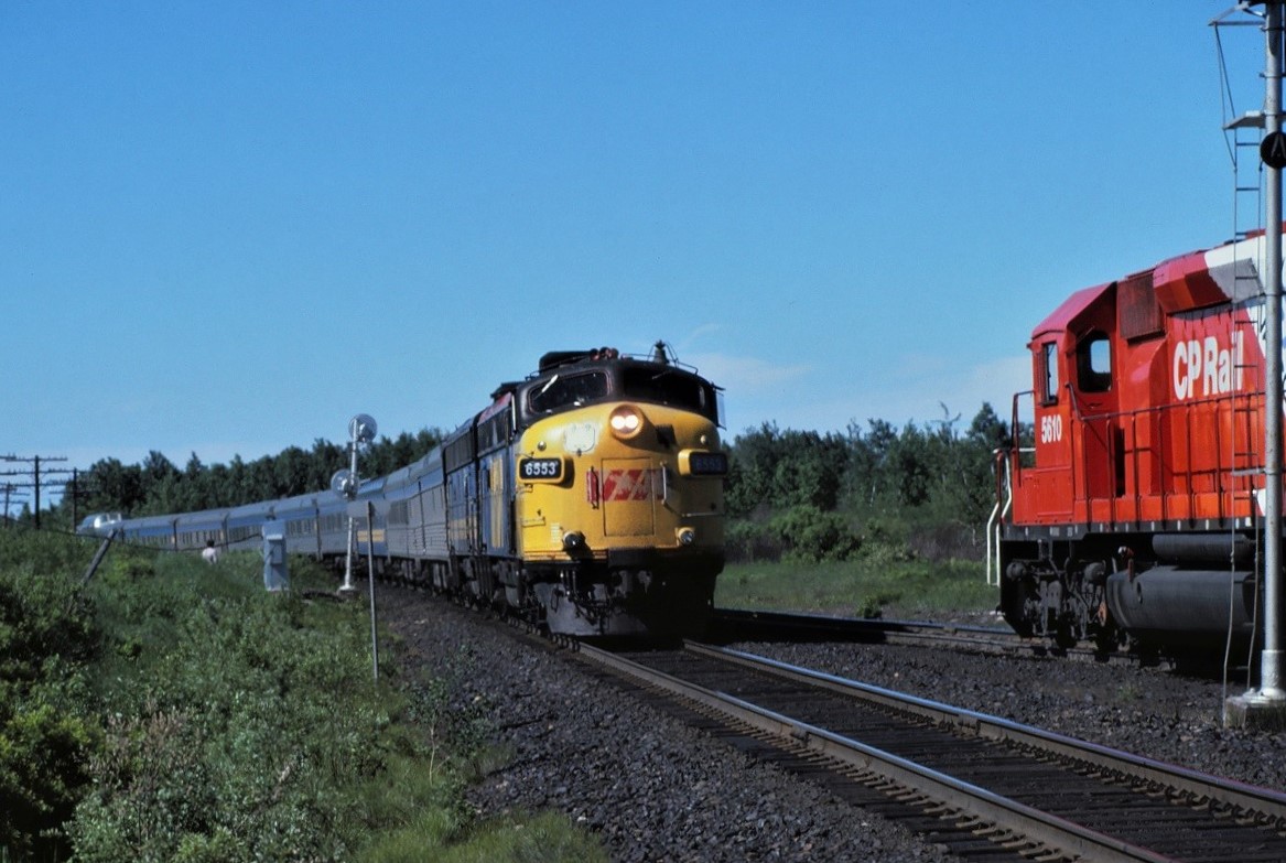 As CP Extra 5610 North sits in the siding at Rutter, Ontario, VIA's southbound Canadian, probably # 10 at the time (my VIA/CP time tables and memory elude me at this time) flies by on the main line. Notice the conductor (brakeman) hiding in the bushes to give the passenger train its roll-by. This track was ABS territory at the time. It was really unusual(to me) to see the freight waiting in the siding looking at a Clear (green) signal; only to have it turn to red (stop) when the passenger train entered the block. Then about 10-12 minutes later the Canadian appears. As soon as it cleared, the signal turned green and the freight proceeded.  Power on the Canadian was 6553 and 6653.