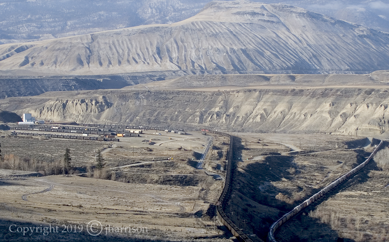 The yard is Ashcroft Terminal, a privately owned transloading and storage facility located a short distance east of the town of Ashcroft, and both trains are westbound. The coal train is on CPs Thompson Sub, and will run through the yard at Ashcroft. The train to the right is a CN on CNs Ashcroft Sub and will skirt the town, as both head for Spences Bridge and beyond. The shadows in the bottom of the image show how sunrise works here at high noon in the fall.