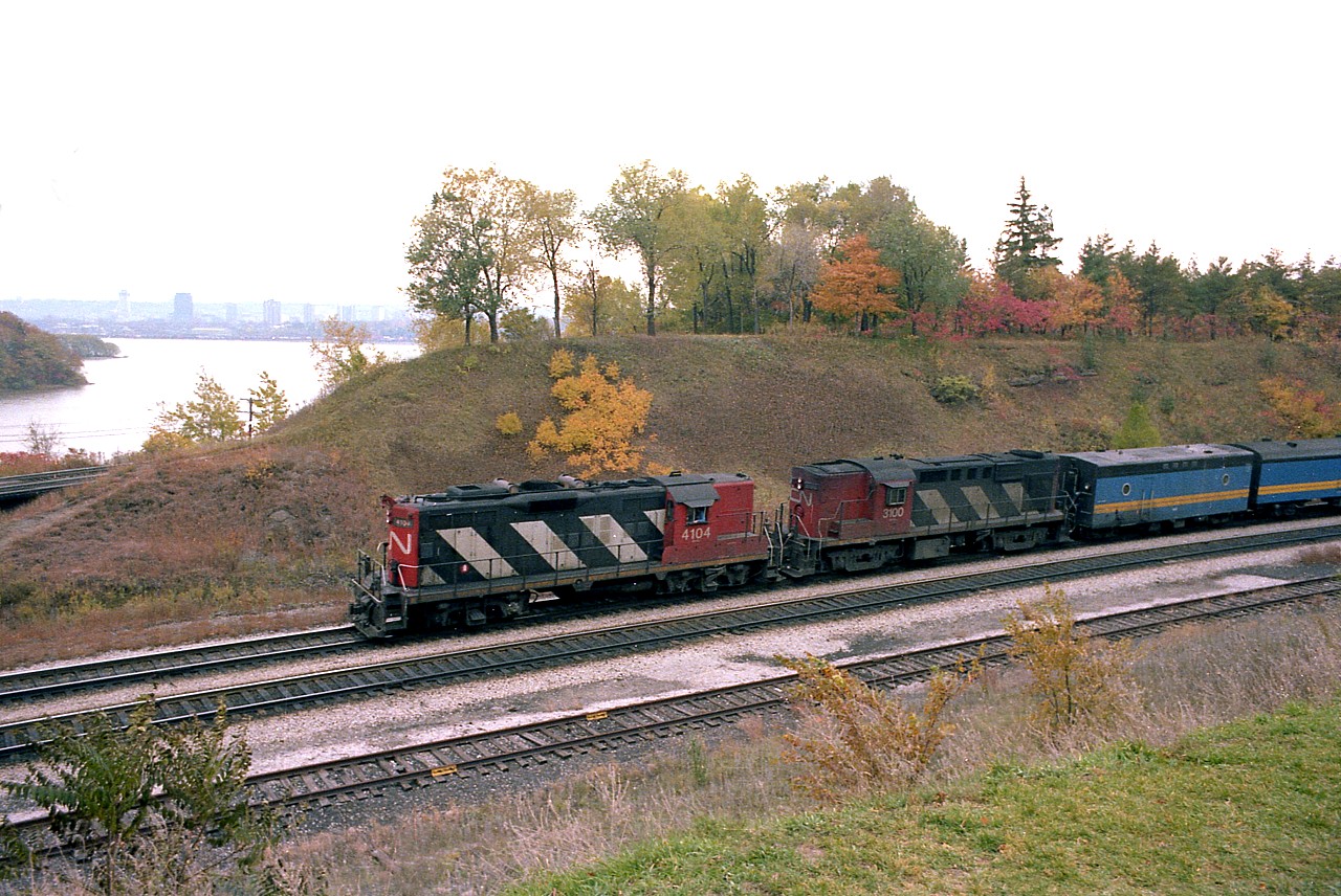 It is late fall but the hillside colours look pretty good. The lands look a bit cleaner as far as bushes and weeds are concerned compared to nowadays.
It is late in the day as VIA #76 approaches the junction running eastward. Some rolling stock is evident in the new VIA colours, but old CN power is still in charge. CN 4104, a GP9 later became CN 4364 and then in 1986 converted to slug #240. Trailing unit CN 3100, an RS-18, was retired from the roster by 1992.
