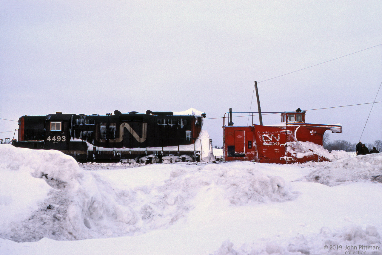 GP9 CN 4493 is powering a plow train, with plow CN 55369 at the head end and looks like another GP9 helping.
The train appears to be halted, with some of the crew on the ground ahead of the plow. 
Could be they are considering their next move. Engineer's seat in the GP9 is occupied.
Location mapped is approximate.
