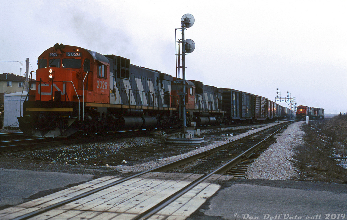 An eastbound freight rolls through Goreway interlocking (and over its namesake road crossing) on CN's Halton Sub with CN C630M's 2026 and 2004 in the lead. In the background, a set of power (M636, M420W and HR616) sits on one of the leads to Brampton Intermodal Terminal, probably waiting to leave for MacMillan Yard for servicing after dropping their train at BIT.  Built in the late 1970's on what was once Chinguacousy Township farmland, Brampton Intermodal Terminal is CN's main GTA-area intermodal terminal and handles traffic going to and coming from Western Canada, Eastern Canada, and the USA. BIT was expanded at various points in time to handle the growning volume of intermodal traffic (although right now it's constrained by development on all sides, and CN is looking at opening another terminal in Milton). The twin leads at the south end connect with CN's Halton Sub at Goreway and Torbram.  The earliest mention of BIT I've found is in some 1975 City of Brampton council minutes. It had been reported as various names before it opened, including "Malport Intermodal Terminal", "Bramalea Intermodal Terminal" (Bramalea amalgamated with Brampton in 1976), "Brampton Intermodal Terminal", and most curious, "Bramport Intermodal Terminal". It appears to have opened as Brampton Intermodal Terminal in 1979 though (for a 1981 aerial view showing how it originally looked, see here (on the left). There was a "ConPort" terminal at MacMillan Yard that likely handled what container and piggyback traffic there was prior to BIT opening (located at the north end of the yard near the old piggyback terminal).  Keith Hansen photo, Dan Dell'Unto collection slide.