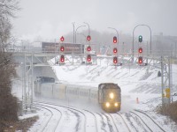 VIA 67 with VIA 6412 up front and VIA 6415 on the rear passes underneath a pair of signal gantries on a snowy afternoon. At right VIA 62 is lined on the south track of CN's Montreal Sub. In the background a Tim Hortons truck takes the ramp for the westbound Autoroute 20, adorned with the slogans 'Always fresh' and 'Toujour frais.'.