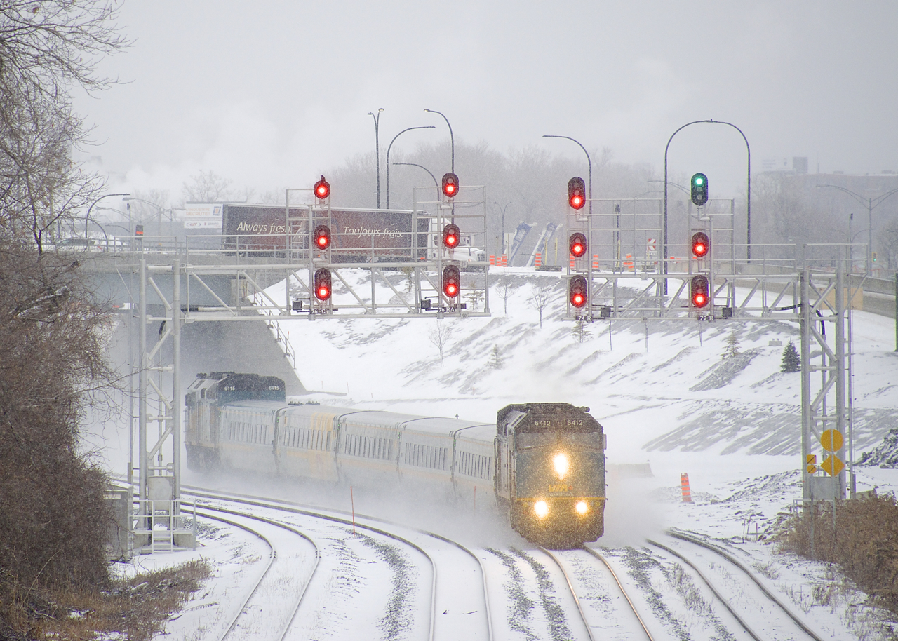 VIA 67 with VIA 6412 up front and VIA 6415 on the rear passes underneath a pair of signal gantries on a snowy afternoon. At right VIA 62 is lined on the south track of CN's Montreal Sub. In the background a Tim Hortons truck takes the ramp for the westbound Autoroute 20, adorned with the slogans 'Always fresh' and 'Toujour frais.'.