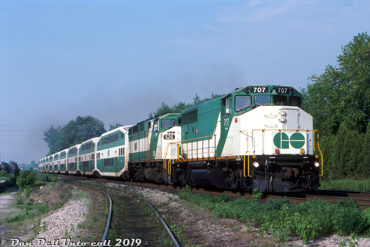 It wasn't very common to see the new GO Transit F59PH units operating with older power, partially because of the short overlap of only a few years that both co-existed on the roster, and partially because the F59's were more than capable of handling a train themselves and providing their own HEP, but it did happen as evident here!

GO Transit GP40-2W 707, running out its final miles on GO just a few months before retirement, double-heads a 10-car bilevel consist with F59PH 526 on train #1908, heading eastbound over Clarkson Road North east of the GO station just before 9 in the morning. The service track to CN's Clarkson Yard, the east end of the wye and Petro Canada's Clarkson refinery can be seen in the foreground.

By this point in time GO had amassed a fleet of 42 F59's in three orders, allowing it to sell both its GP40TC's (to Amtrak in 1988) and F40PH's (to Amtrak in 1990). Their GP40-2W and rebuilt GP40-3's remained on the roster, often leased out to CP for freight service, but around the fall of 1991 ten GP40-2W's including 707 would be sold to CN for freight use and renumbered above their own 9600-series units (a lone unit, 703, was sold to Tri-Rail). 

The ex-Rock Island GP40-3's would solder on for a few more years (often being leased to power-short CP for freight service) before being traded in to GMD on the last order of F59's in 1994, giving GO a solid roster of 49 F59PH units. Aside from a sale of 4 surplus units to TRE in 1997, the fleet of F59's would remain as GO's sole power until the first new MP40 arrived in the fall of 2007.

Peter Jobe photo, Dan Dell'Unto collection.