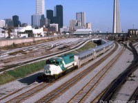 Here's some GO 708 action to go with Steve's shot of <a href=http://www.railpictures.ca/?attachment_id=39978><b>CN 9675</b></a>: GO Transit GP40-2W 708 heads up a short consist of five Hawker Siddeley single levels and an APCU, heading westbound out of Toronto Terminals Railway territory at Bathurst Street bridge. 708 was part of GO's final GP40-2W order, units <a href=http://www.railpictures.ca/?attachment_id=35761><b>707-710</b></a> built in 1975, continuing GO's trend at the time of ordering freight power to run with power cars in passenger service. In 1991, 708 and most of the GO -2W fleet would become CN 9600's. <br><br> Following the busy and colourful <a href=http://www.railpictures.ca/?attachment_id=15641><b>50's</b></a>, <a href=http://www.railpictures.ca/?attachment_id=36430><b>60's</b></a> and <a href=http://www.railpictures.ca/?attachment_id=30123><b>70's</b></a> eras, I suppose one could call this the bland or malaise era of Bathurst Street: freight operations are gone as CN has finally pulled out of their old Bathurst Street freight yard, and GO has taken over the rest for storing commuter trains. The old CN yard tower that was here is gone. The new concrete "flyunder" has been built tracks haven't been laid yet (note the fresh-looking re-aligned mainline tracks in the foreground to make way for its construction, as well as the yard leads on the left being reconfigured). The downtown skyline has a healthy growth of office towers, but still looks plain and uncluttered compared to today's condo and high-rise boom. The colourful mix of red CP and black/white/red CN passenger trains have been merged into the blue and yellow of VIA. And the old CN/VIA Spadina roundhouse and coachyards remain, but for only a few more years until VIA moves all operations to Mimico in the mid-80's to clear room for the Skydome. <br><br> Things didn't improve too much by the <a href=http://www.railpictures.ca/?attachment_id=4210><b>mid-2000's</b></a> (much of the coachyard lands to the south were just starting to see development), but by the <a href=http://www.railpictures.ca/?attachment_id=6182><b>2010's</b></a> condos started popping up everywhere downtown. <br><br> <i>Robert D. McMann photo, Dan Dell'Unto collection slide</i>.