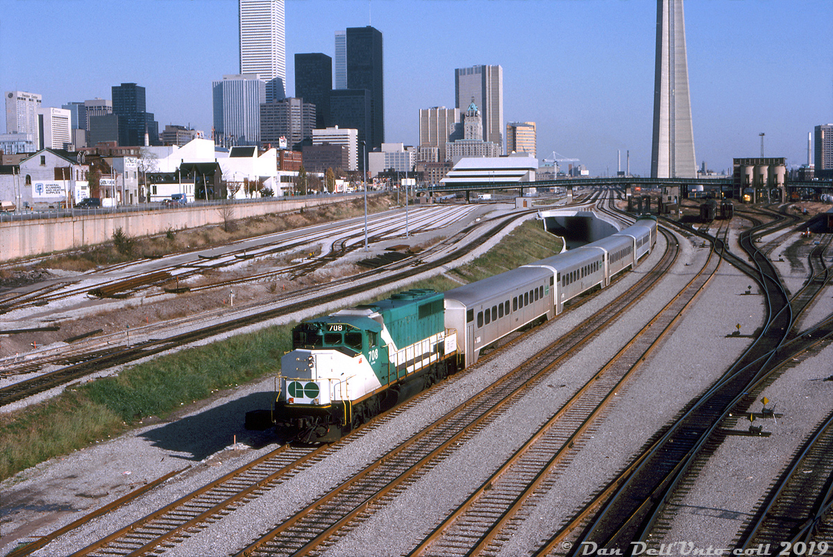 Here's some GO 708 action to go with Steve's shot of CN 9675: GO Transit GP40-2W 708 heads up a short consist of five Hawker Siddeley single levels and an APCU, heading westbound out of Toronto Terminals Railway territory at Bathurst Street bridge. 708 was part of GO's final GP40-2W order, units 707-710 built in 1975, continuing GO's trend at the time of ordering freight power to run with power cars in passenger service. In 1991, 708 and most of the GO -2W fleet would become CN 9600's.  Following the busy and colourful 50's, 60's and 70's eras, I suppose one could call this the bland or malaise era of Bathurst Street: freight operations are gone as CN has finally pulled out of their old Bathurst Street freight yard, and GO has taken over the rest for storing commuter trains. The old CN yard tower that was here is gone. The new concrete "flyunder" has been built tracks haven't been laid yet (note the fresh-looking re-aligned mainline tracks in the foreground to make way for its construction, as well as the yard leads on the left being reconfigured). The downtown skyline has a healthy growth of office towers, but still looks plain and uncluttered compared to today's condo and high-rise boom. And the old CN/VIA Spadina roundhouse and coachyards remain, but for only a few more years until VIA moves all operations to Mimico in the mid-80's to clear room for the Skydome.  Things didn't improve too much by the mid-2000's (much of the coachyard lands to the south were just starting to see development), but by the 2010's condos started popping up everywhere downtown.  Robert D. McMann photo, Dan Dell'Unto collection slide.