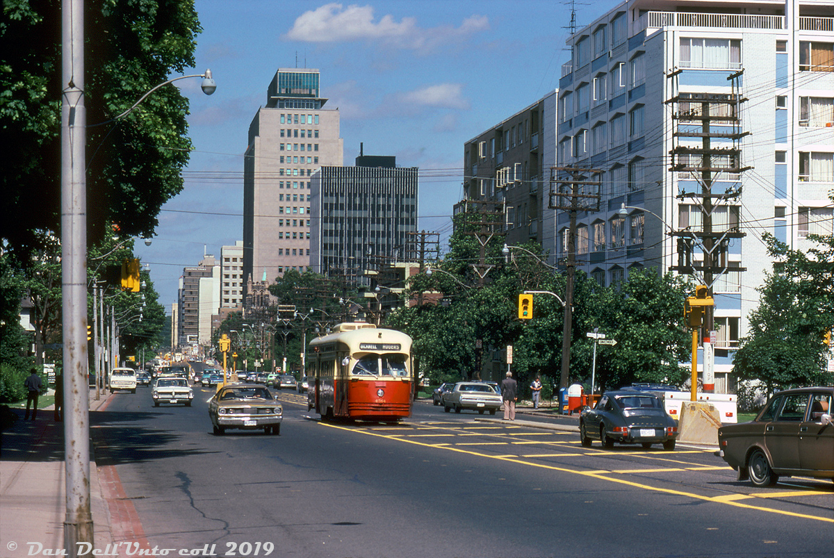 TTC PCC 4561 (an A9-class streetcar originally bought by Cincinnati in 1947) heads west in traffic through Midtown Toronto on St. Clair Avenue, operating on a westbound/northbound Rogers Road streetcar bound for Bicknell Loop. The office buildings and highrises around Yonge Street are visible in the background. To the north (left) is the upscale Forest Hill area full of posh housing, the smell of old money, and prominent families of wealth such as the Eatons. The two individuals pictured on the left may have been fellow transit enthusiasts, as this was during a special 3-day series of fantrips held on July 5-6-7th. 

Normal routing for the Rogers Road streetcars was between Oakwood Loop (Oakwood/St. Clair) and Bicknell Loop (Rogers/Bicknell), but rush hour cars were extended to the subway and ran along this stretch to/from St. Clair Subway Station on the Yonge line. By the time of this photo, there was less than a month left for the Rogers Road streetcar: operations would end after July 19th 1974 and be replaced with a branch of the Rt.63 Ossington trolleybus route.

Robert D. McMann photo, Dan Dell'Unto collection slide.