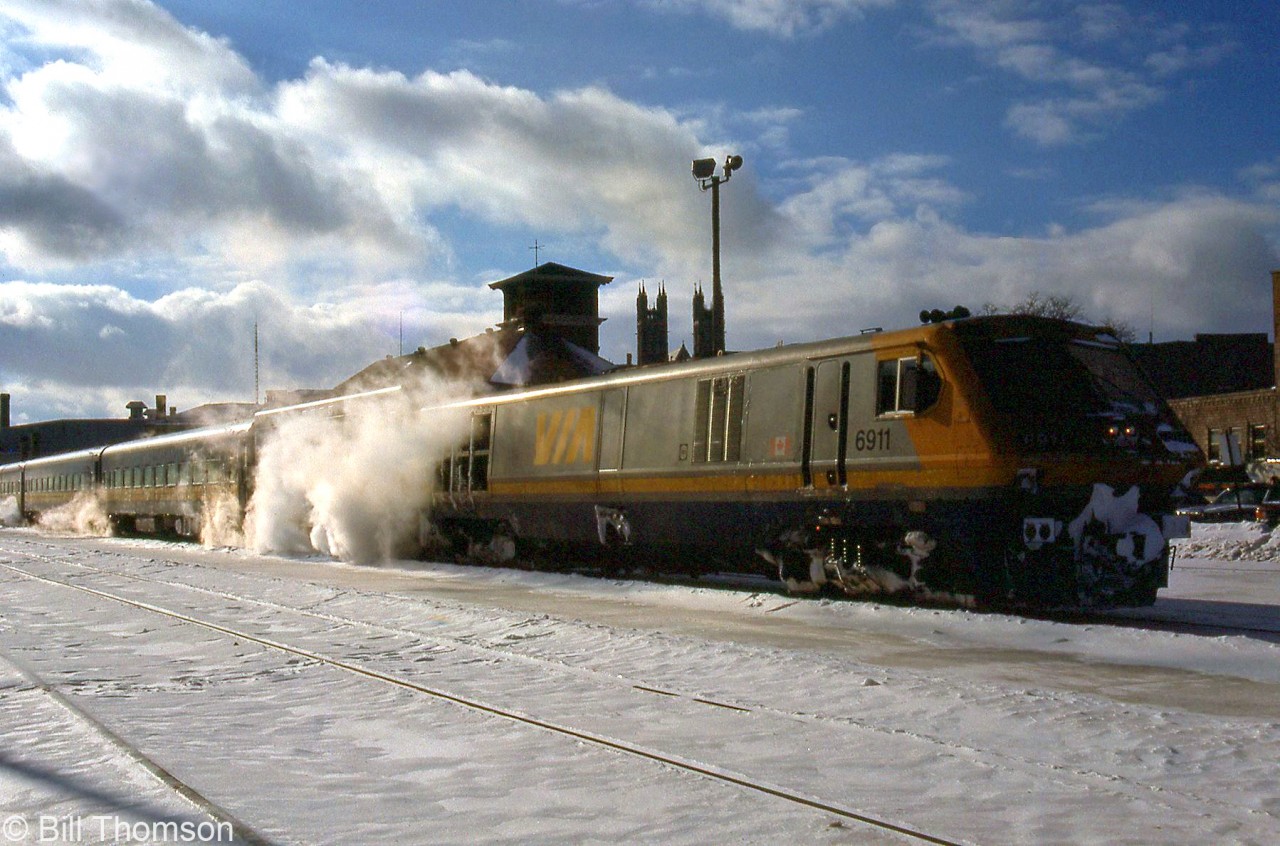 VIA LRC 6911 leads a steam-heated passenger consist of blue and yellow cars on a chilly day in January 1990, stopped at the station in Guelph. Steam generator cars were necessary when running older equipment with LRC or F40PH locomotives, as they were delivered with HEP generators for modern equipment, rather than steam generators that the older cars required.