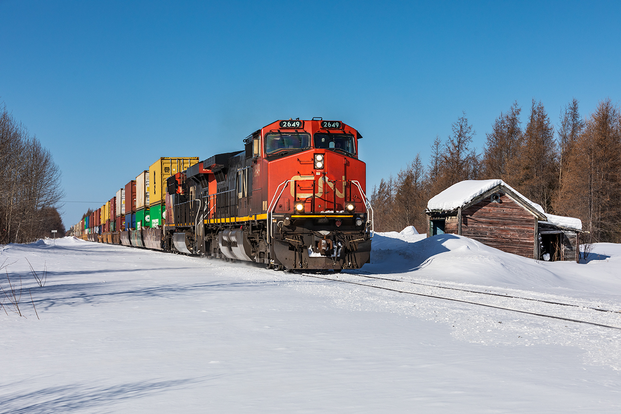 Twenty-four hours before this photo was taken, I was sitting in the waiting room of the Toyota Dealership in North Bay. Twiddling my thumbs anxiously as Toyota assessed the damage that a three-inch steel spike left in my tire the night prior - stranding me in town for the night. Things were looking grim. Discouraged by sinking hundreds of dollars into the whole blow out ordeal, I questioned if it was worth continuing on. But that voice inside of my head reminded me that this was, likely, a once-in-a-lifetime opportunity and to go for it. So, I pressed onwards. Following the road north to Englehart, it was a cloudless, blue-sky day. The next morning was much the same as I woke up at sunrise and followed 'Ontario's Development Road' to Cochrane where Canadian National was operating a detour stack train over ONR's Kapuskasing Subdivision. Normally relegated to four-axle power, it came as quite a surprise to area-railfans when CN started operating their big stack trains over the rickety Hearst to Cochrane line. A last resort, as a derailment in Haileybury put ONR's Temagami Subdivision out of comission for a number of days. 

If there was one shot along the Kap Sub that I wanted - aside from the station at Cochrane - it was at the former National Transcontinal Railway sectionhouse at Driftwood. So, I put on my snowshoes (a must as the snow is 3 to 4 feet deep in most places) and trekked into the woods. Moments later, a headlight appeared on the horizon, followed by the most surreal sight of double-stacked containers. Here she comes! Rattling snow off of the steam-era NTR sectionhouse, CN 2649 rolls along ONR's Kapuskasing Subdivision with detoured stack train no. 188 at Driftwood, ON. In 17 miles, 188 will reach the southern terminus of the Kapuskasing Sub at Cochrane, and change crews at the beautifully constrcuted depot. 

After the North Bay chagrin, I'll consider this photo a consoulation prize.