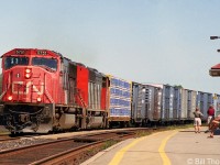 CN SD75I 5751 leads an SD60F on a westbound freight, passing by passengers waiting at the VIA station in Cobourg on a sunny afternoon.