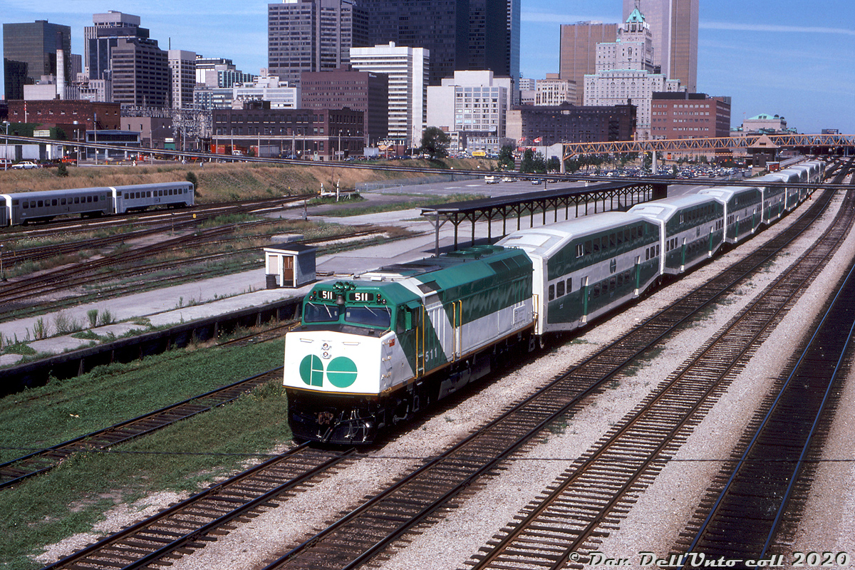 Only a few months old at the time, shiny GO Transit F40PH 511 heads up a westbound 9-car train of new bilevels and an APCU on the tail end, passing by Spadina Avenue just west of Toronto's Union Station. GO 511 was part of a six-unit order of F40PH units built by GMD at their London ON plant in April-May 1978, a change from GO's previous orders of freight GP40-2W units. New F59PH deliveries a decade later would result in GO flipping the six orphan units to Amtrak. 511 would see another decade of service on Amtrak as their 411, and would go on to work AMT's commuter operations out of Montreal. It last saw service on the the Saratoga & North Creek Railway (via LTEX) with another ex-Amtrak F40.

The bilevel cars trailing are also new, part of the first order built by Hawker Siddeley and in service for a little over a year since introduction in March 1977. Since the first bilevel order included no cab cars, another locomotive, APCU, or old Hawker Siddeley single-level cab car had to be used on the other end in the early years.

This scene off Spadina Avenue is notable in that it's under transition: the old CN steam-era freight platforms, team tracks and yard trackage east of Bathurst Street yard are still present, but truncated for the new CN Tower's parking lot (note the yellow bridge crossing the rail corridor near TTR's John St. interlocking tower). In later years the RBC's new data center (325 Front St. W), the Metro Toronto Convention Centre and CN's L'Hotel would fill much of the vacant land along Front Street in the 80's. The usual downtown office towers of the day including the TD Centre, Royal Bank Plaza, CP Hotel's Royal York, and the CNCP Telecommunications Building (151 Front Street) are quite unobscured compared to today.

Bill Mischler photo, Dan Dell'Unto collection slide.
