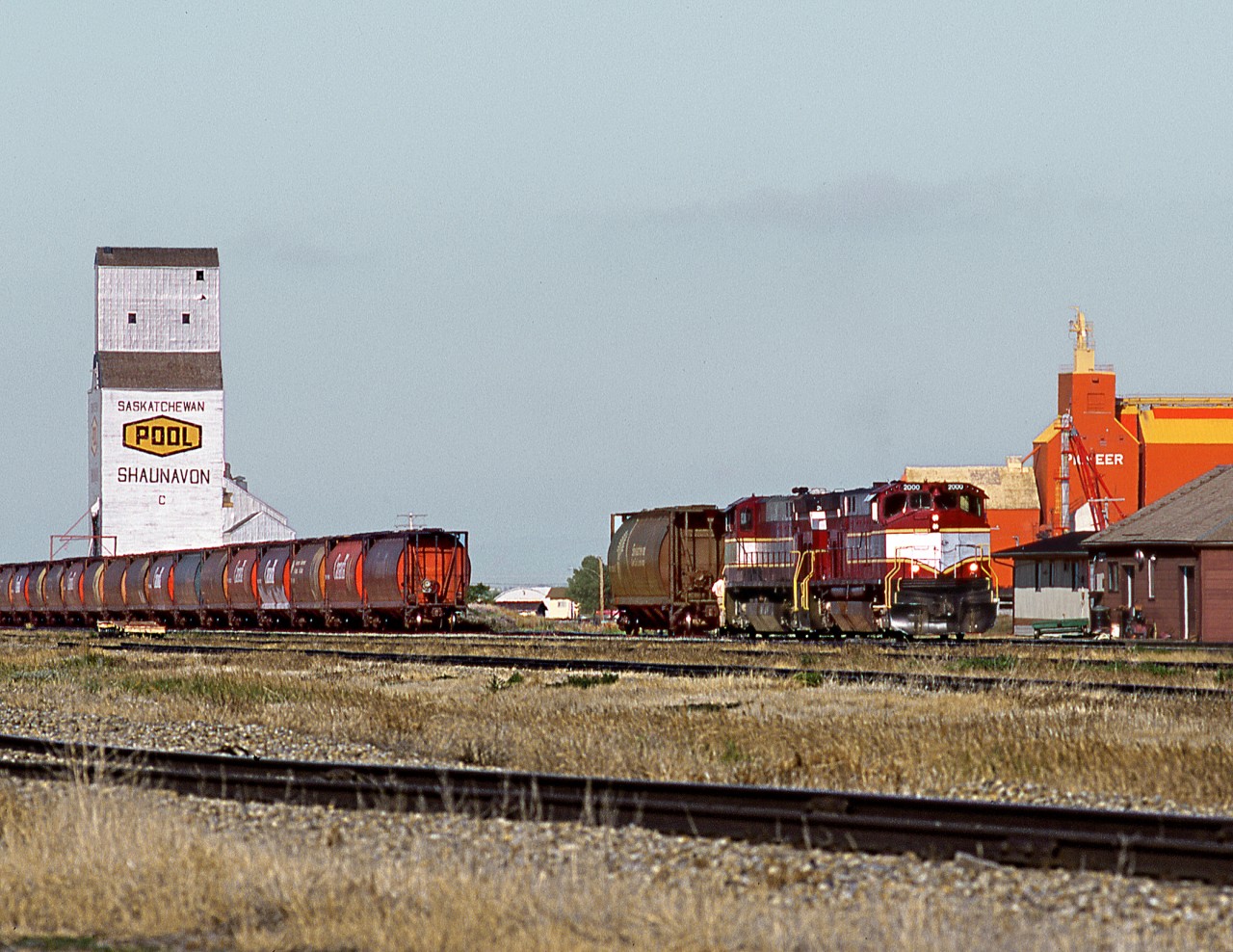 Great Western's M420's still in Bay Colony paint scheme make up a train of grain loads for delivery to CP at Assiniboia. The Atco trailer near the unit is company HQ and the wooden building is tool shed, which was freight shed of CP's now demolished 3 story station