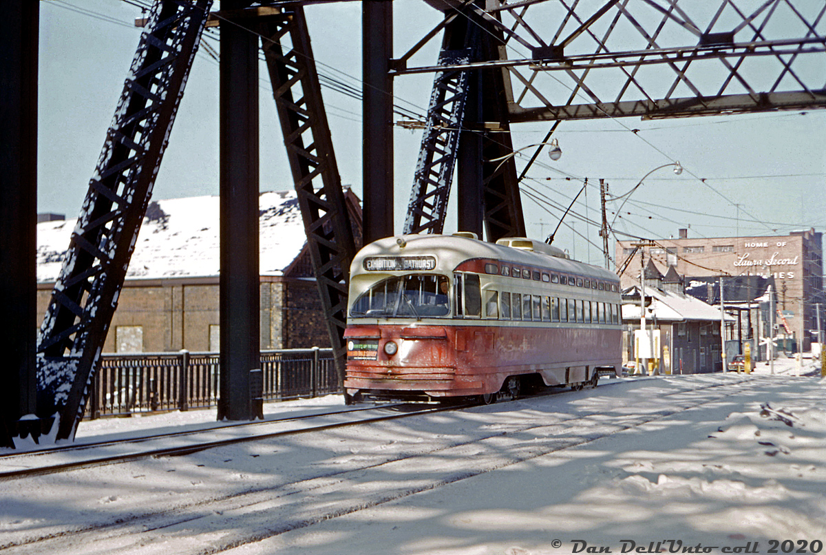 In another shot of the Bathurst Street bridge reconstruction work in 1963-1964, TTC PCC 4393 (A6-class, built 1947-48) heads southbound on Bathurst Street south of Front, operating on temporary track laid on the west side of the bridge for southbound streetcars (the actual southbound track in the foreground was being used for northbound streetcars, while the two right lanes were closed and being redone). Note the relatively undisturbed snow on the bridge, as it was only open for streetcars and closed to regular automobile traffic.

The property to the northwest of the bridge behind the streetcar, once home to the Consumers Gas Company (one of their old coal gas manufacturing facilities, that had two very large gasometer tanks on site), was now home of the Kaufman Metal Company and the Toronto Refiners & Smelters Co. Visible in the distance is the Laura Secord candies building around Wellington. A keen eye would pick out something out of place here: a set of railway crossbucks. Those were for a industrial spur that lead across Bathurst Street (and the streetcar tracks at-grade via diamonds) to get to the Bathurst Tool building on the east side of Bathurst north of Front. As far as I can tell, this spur fell out of use sometime in the 1950's and was removed in the early-mid 60's, although I haven't found any decent shots of streetcars crossing it. For a 1947 aerial showing the crossing, click here (check near the middle of the image).

Photographer unknown, Dan Dell'Unto collection slide.