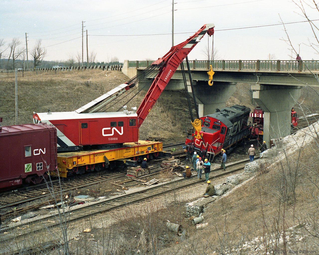 GP9rm mother unit CN 7263 will have its front lifted by Toronto Auxiliary crane CN 50008 to get it back on the rails. The paint on CN 7263 is clean and glossy, so it's not long since its 1990 remanufacture.
CN 7263 had been in a collision with GP38-2 hump switching unit CN 7508 which appeared to have got the worst of it, including having all its right side hood access doors torn off. Both units returned to service after repairs.
According to my 1990 Trackside Guide, CN 50008 was a 250 ton capacity Bucyrus-Erie built in 1946. 
Note the outriggers extended from the crane base and woodblock piles beneath to counter tipping forces.
The bridge carries Rutherford Road over Macmillan Yard hump pullback tracks.
Alan recalls taking these pictures in 1991. Foliage colour and some residual snow suggest March.