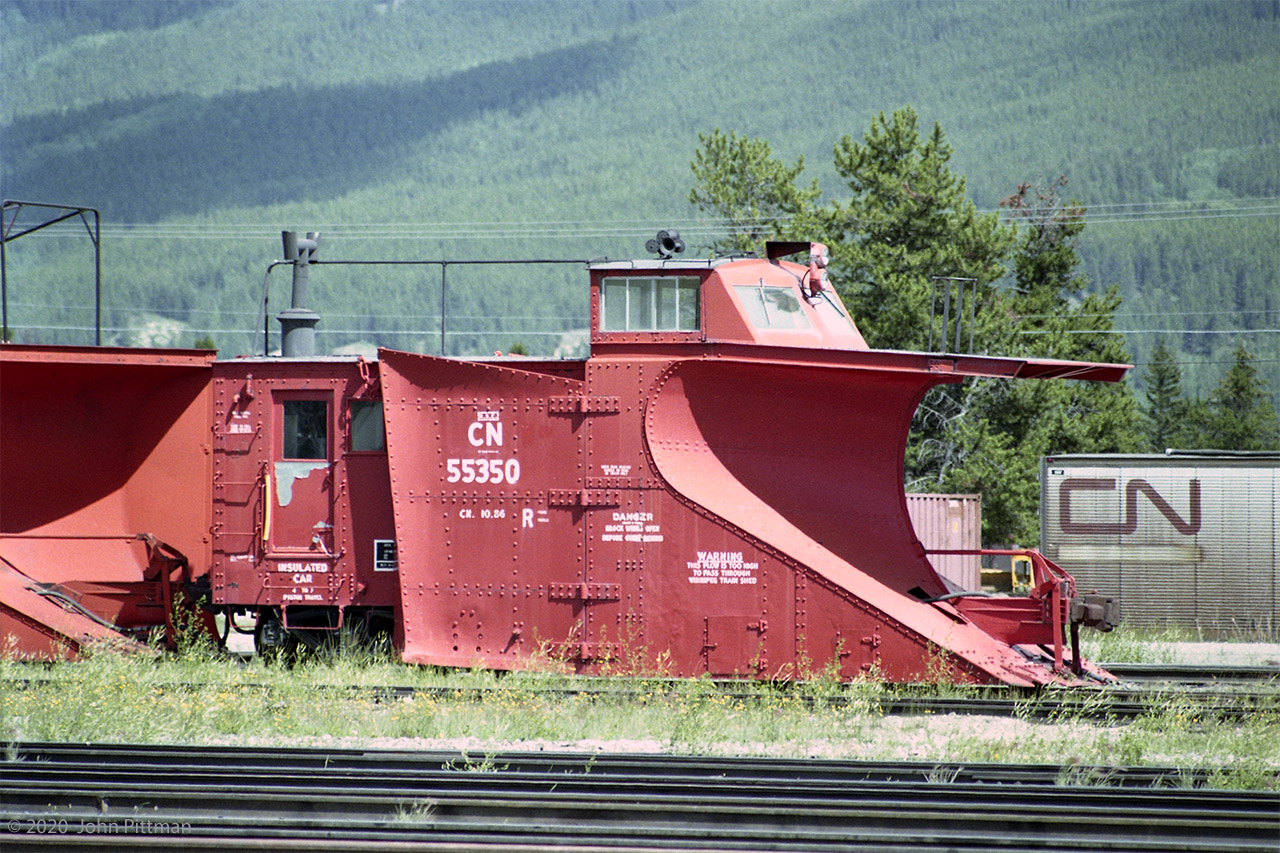 Single-track snowplow CN 55350 is in front of a dual/multi-track unit that moves plowed snow to the right.
According to my Trackside Guide, CN 55350 was built in 1928 by the Eastern Car Company of Truro NS.
Lettered on the side is "CN 10.86" - could be date of rebuild or major maintenance - and some notices: 
"DANGER - Block Wings Open Before Going Behind" 
"WARNING - This Plow Is Too High To Pass Through Winnipeg Train Shed"
It's a colourful summer scene, but ... Winter is coming.