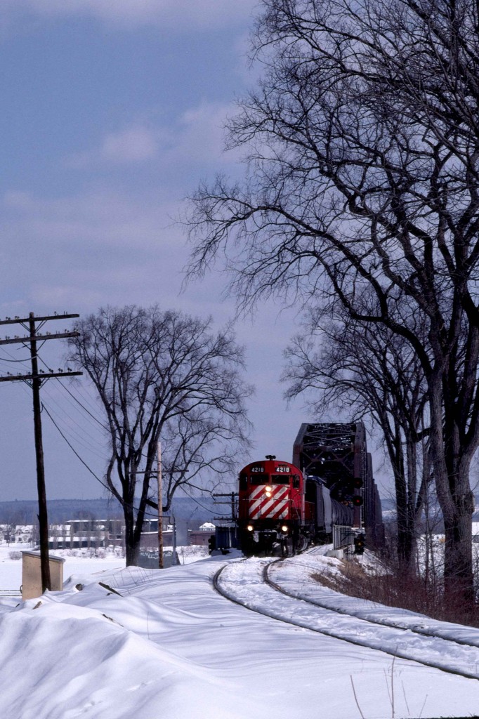 The Nackawic Turn is close to its home terminal in Fredericton, New Brunswick, on a beautiful afternoon in March 1985.  It is about to pass the site of the former CNR Fredericton station on the engineer's side of the tracks.  

C-424 4218 was built in Montreal, Quebec, by MLW and delivered as the 8318 on Tuesday, October 5, 1965, with serial 84856.  The CPR received trade-in credit with the return of Alco FB-1 4402 on Thursday, July 8.  The unit was part of 32 unit order 4905, class DRF-24b, built between March and December 1965.  Their delivery was followed immediately by order 4906 for 18 units of class DRF-24c.  MLW built these without trade-ins from December 1965 through March 1966.  CPR initially used the C-424s on priority freights, notably on the Eastern Region, as St. Luc was their original maintenance assignment.  When they were new, I often saw them on Windsor - Montreal hotshots in combination with the nearly new GP35s, which had proven slippery.  CPR quickly renumbered the units in the 4200 series.  Retirement, conversion or sale came to the C-424s before 1996; however, the 4218 was not one of the 31 survivors.  

The Nackawic Turn left Fredericton weekday mornings with traffic for the paper mill at Nackawic on the north side of the Saint John River.  At Una Junction, Mileage 21.8 of the Fredericton Sub, the outbound train turned north on trackage rights at Mileage 0.2 of the CNR Fredericton Spur and joined their 70.5-mile Saint John - South Devon Oromocto Sub at Mileage 69.4.  The Turn then crossed the bridge in the background.  The ice blocked river destroyed the original crossing; this bridge opened in 1936.  (It survives as a walking path following the end of rail service in the late 1990s.)   Reaching South Devon on the northern bank, the train turned north for a 37-mile run to Southampton on the Gibson Sub.  At Southampton, Mileage 22, it turned south, travelling 9.4 miles to the mill at Nackawic.   

Upon returning to Fredericton, the power was set up to do a nightly 133.2-mile turn to Saint John.  This arrangement ended on Friday, October 25, 1985, thereafter power from mainline freights brought cars the 22.2 miles into town from Fredericton Junction on the Montreal - Saint John line.   Prior to Friday, November 28, 1980, the nightly freight, the "Oriental', took its train to McAdam, forty miles west of the Junction.  

The 1967 flooding behind the Mactaquac dam caused the relocation of several communities. It spawned the province's first model community in Nackawic.  The flooding also cut short the last 3.1 miles of the Southampton Sub that reached down to the Saint John River at Otis.  St. Anne Nackawic Pulp and Paper opened their kraft pulping mill in 1970 and received a continuing supply of rail-hauled Bunker 'C' oil from the Irving refinery in Saint John.   Rail shipments lasted until December 1994 when CP Rail discontinued its operations east of St. Jean, Quebec and abandoned its branchlines north of Fredericton.   The present owner, AV Group NB Inc., produces up to 540 metric tons of bleach kraft pulp daily.