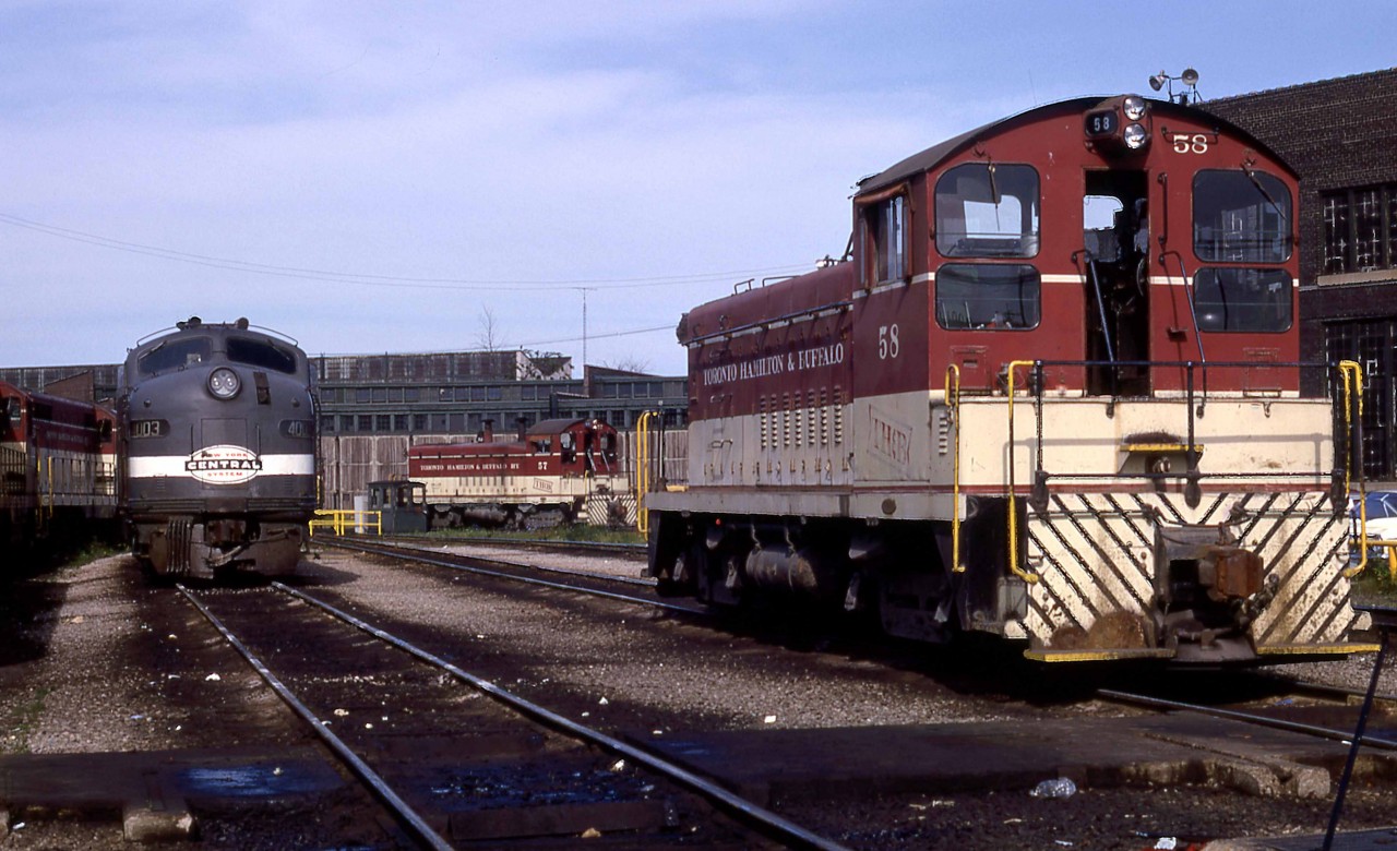 Toronto Hamilton & Buffalo SW9 58 rests near the railway's Aberdeen Roundhouse in Hamilton, Ontario, on Sunday, July 7, 1968.  GMDL built the 1200hp switcher in nearby London, Ontario, in January 1951, serial A-124, as their 25th production unit.  The 58 was equipped with mu and was the last of four ordered by the TH&B in 1950 to complete dieselization of their switching services.  Retired in May 1988 and sold to Atlas Steel in Welland, it was resold that year to Lukens Steel's Brandywine Valley Railroad in Coatesville, Pennsylvania, as their first 8206.  It was transferred to the Upper Merion and Plymouth at Conshohocken Pennsylvania in 1991, where it continues to operate as their second 9007.    

Facing the switcher is NYC E8A 4003 fresh from an early morning run from Buffalo on Toronto-bound Penn Central Train 371.  Connecting Train 57 had departed Grand Central Terminal the previous evening with coaches and sleepers for Toronto.    My friend, Bruce Mercer, was on duty at Port Credit that morning and reports that CP RS10 8575 handled the last 39.3 miles from Hamilton to Toronto as Train 322.  

New York Central 4003 was built as an E7A at LaGrange in March 1945 with serial 2868.  It was involved in a wreck at Conneaut, Ohio, on March 27, 1953, and was rebuilt by EMD as an E8A.  Not long after, it was on the September 1956 cover of Trains Magazine in an image by Phil Hastings and featured in the particularly famous story by David Morgan: The Mohawk That Refused to Abdicate.   The unit was retired in June 1973.  

The TH&B completed its line between Waterford and Welland, Ontario, via Hamilton in 1895.  NYC (73%) and the CPR (27%) purchased the railway in 1895.   In 1977 CP Rail bought the former NYC shares and merged it entirely in 1987.