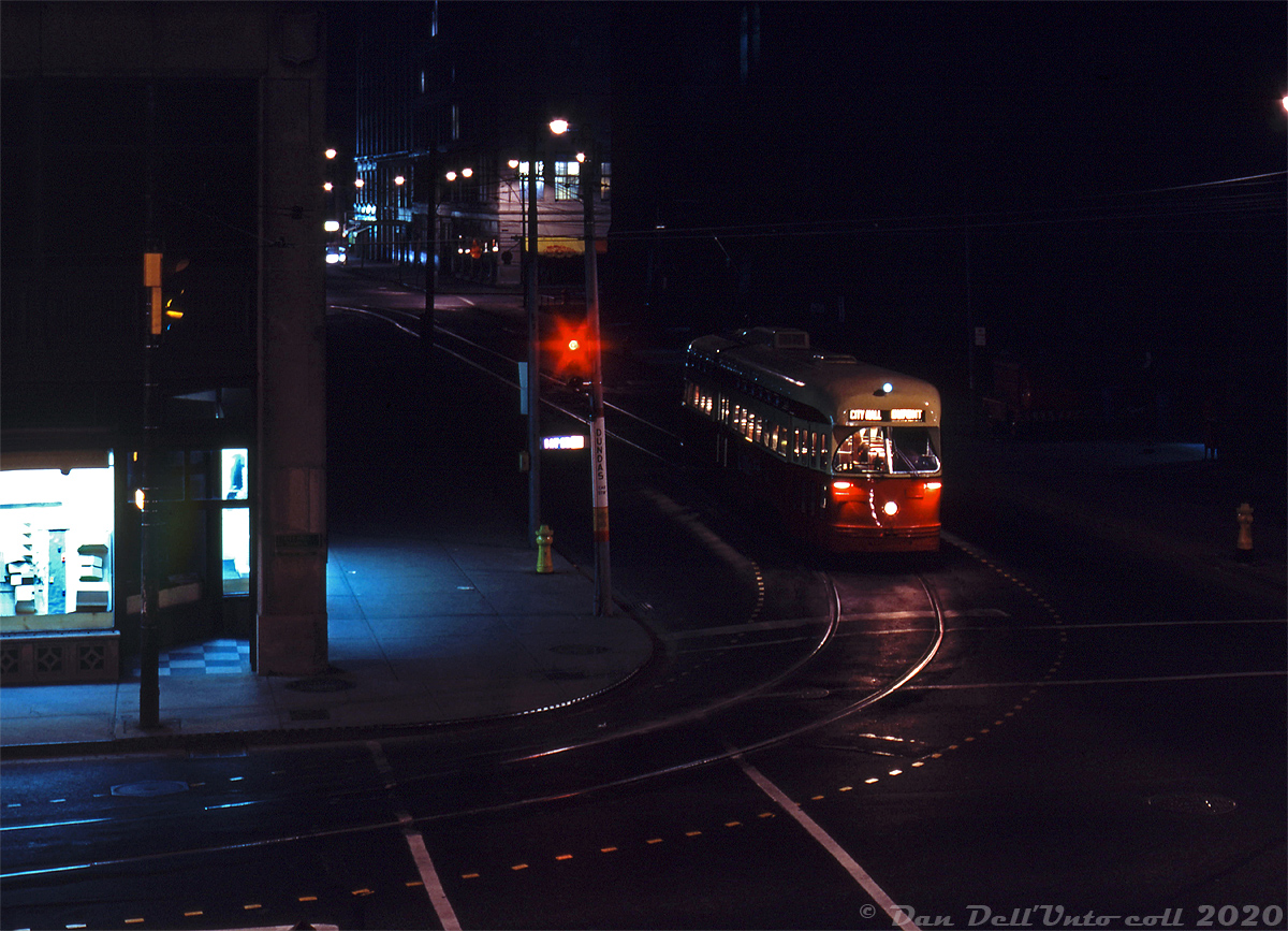Night Owl Trip: Operating on an all-night fantrip, TTC PCC 4732 (one of the 48 1947 Pullman-built A13-class cars acquired secondhand from Birmingham, Alabama) poses for a night-time photo stop at the corner of Bay Street and Albert Street in downtown Toronto. The car is turning on City Hall loop, an on-street loop of track off Dundas running clockwise from Louisa-James-Albert. Lurking in the shadows of early 1970's street lighting are the Old City Hall (dark, to the right), Eaton's Annex store (to the left), and Eaton's flagship Queen Street Store (in the distance at Albert & James). Note that Albert Street still ran all the way from Bay to Yonge Street at the time.

It had been common since the 1960's for railway societies and individuals (both local and visiting) to charter streetcars for private trips touring around the city, taking photos, and to visit different transit carhouses, garages, terminals, loops, etc along the way. Standard practice during a photo stop was to sign a car up for some of the routes that operated in the area, past and present. In this case, 4732 has been signed up as a Dupont car bound for City Hall (loop), even though the Dupont route vanished almost a decade earlier in 1963 when the University subway line opened (see the Bill Thomson's photo here).

In the coming years, the new Eaton Centre development would close some of the local streets and eliminate City Hall loop. The Eaton's Queen St. store would get demolished to make way for further mall expansion (once the new flagship Eatons store at Dundas was opened), and Albert Street would be truncated at James. 

Robert D. McMann photo, Dan Dell'Unto collection slide.