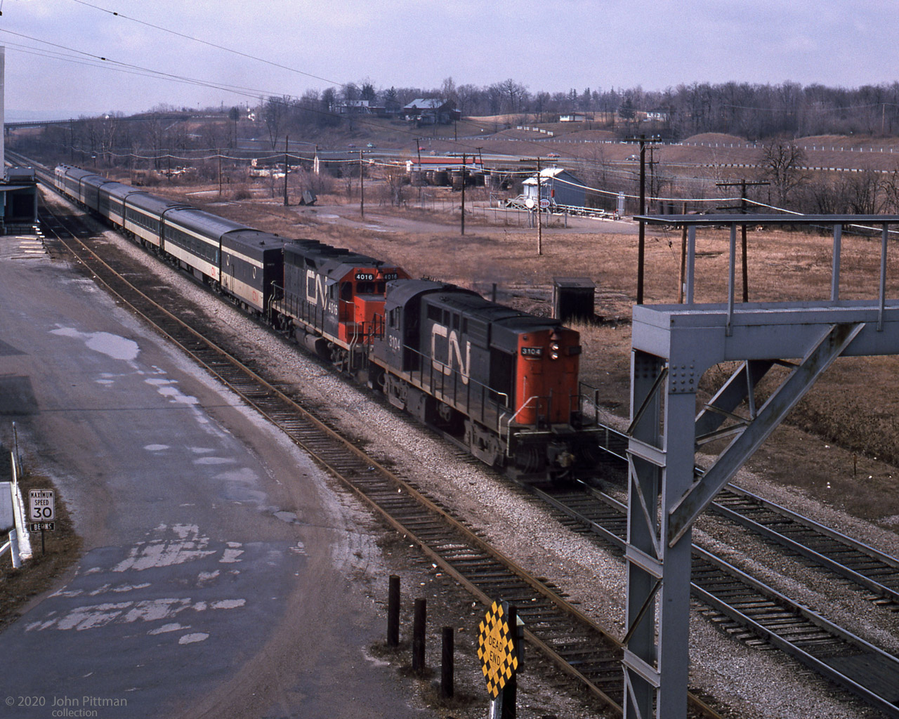 A CN morning passenger train speeds eastward past Aldershot Cold Storage, about to pass under Waterdown Road.
Powered by MLW RS-18 CN 3104 and GMD GP40 CN 4016, the train has a Steam Generator unit, 5 coaches, and looks like a baggage car trailing.
The businesses between the tracks and Highway 403, the nearest with Esso signs, are long gone. 
Perhaps they were related to the Cold Storage operation; I wonder how that area used to be accessed.
The major roads and railway remain, but much has changed since 1975.