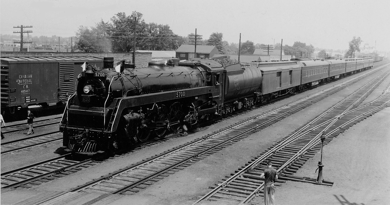 CNR 5700, one of only five K-5-a Hudson locomotives built for the CNR by MLW in 1930 for high speed passenger service in the Montreal - Windsor corridor.  She is seen here leading a passenger extra westbound through Hamilton, likely just east of Ottawa Street based on the house/building at centre, and the distance to the Dofasco structure in the distance.  Date is approximate based on the repainted stencil on the boxcar (CNR 485147) at left.  This car was part of a 1,350 car order of 40 foot boxcars placed by the CNR to National Steel Car in October 1943, the order being built between February and May 1944.  With the weight information restenciled on the side as 5-52, it is possible the car was involved in an accident or altered and reweighted due to the additional structure.At the close of the steam era in 1960, CNR marked 5700 for preservation, however it was unfortunately scrapped by mistake.  Instead, CNR 5703 was pulled from the scrap lines and renumbered as 5700, now on display at the Elgin County Railway Museum in St. Thomas.  The difference between the two 5700's is see on the driving wheels.  All K-5-a locomotives received spoked driving wheels save for class leader 5700, which had boxpok wheels.  One other K-5-a Hudson, CNR 5702, is preserved by Exporail in Saint Constant, Quebec.