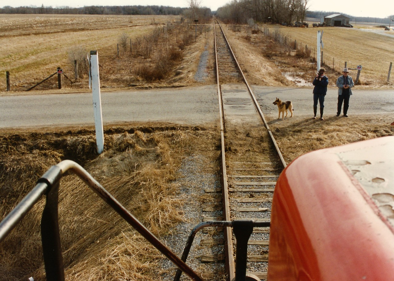 A view from the cab of CN 4115 as it powers the rail salvage train towards Line 60, which was the first crossing west of Palmerston on the CN Newton Subdivision. As well, a couple onlookers and a companion have gathered at the small crossing to take in the historic action. Earlier that day, the rail train had slowly rolled through the town of Palmerston for the last time and was powered by CN GP9RM’s 4115 and 4140. During April 1996, CN had removed the last sections of the Newton and Owen Sound Subdivisions that remained between Harriston and Stratford. Note- Photo taken with permission of the rail train crew.