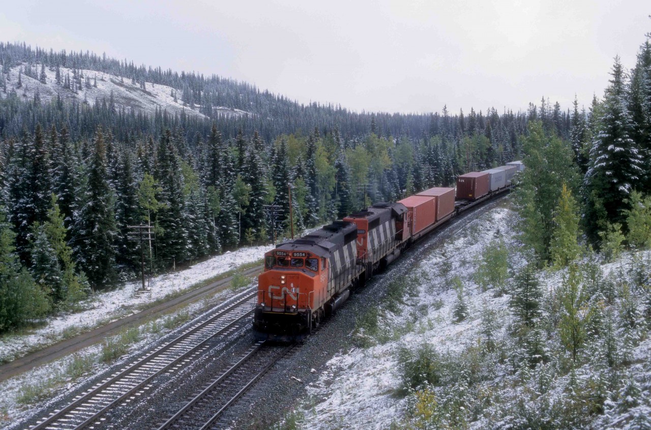 A CN westbound pulls down the Toronto - Vancouver mainline while awaiting an eastbound to pull up the siding at Mileage 190 of the Edson Sub at Entrance Alberta in September 1997.  The train had just passed Hinton and is continuing up the valley of the Athabaska River towards the next division point of Jasper.   Entrance derives its name from the fact that it is an eastern gateway to Jasper National Park in the Canadian Rocky Mountains.  

GP40-2L(W) 9554 (3-1975 #A3189) is one of 268 ordered new from GMDL in London, Ontario, with deliveries of four orders between March 1974 and May 1976.  CN acquired a further ten from GO Train in 1991.  CN's Research Centre developed a Positive Traction Control system that GMDL adopted and introduced as a factory installation on these units.  The new system reduced wheel-slippage, which enabled them to accelerate high-priority, high-speed trains more quickly.   The units traversed the system and became a mainstay on Toronto - Edmonton trains because their lightened and raised frames allowed the installation of 3,000 imperial gallon fuel tanks.  The larger capacity eliminated refuelling between Toronto and Winnipeg.   GMDL installed standard, 2,300-gallon tanks on the final 35-unit order, which are GP40-2s.    

CN's Research developed improvements to the lateral clearance on the centre axles of C-C trucked locomotives beginning in 1975 so that the superior tracking abilities of the GP40-2s became redundant.   Their higher fuel consumption as compared to newer units, led to their early retirement from CN.  Today only 59 of the 278 units remain, mostly in yard and local service.  

9554 left the roster in 2000, and it became Yadkin Valley 9554 in North Carolina and later RLHH 3049 in Southern Ontario.   In March 2019, RLHH traded it in on a GP38-2 from Metro East Industries in Fairview Heights, Illinois, a suburb of St. Louis, Missouri.