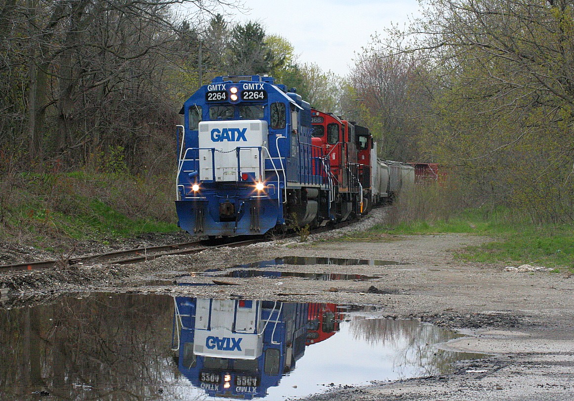 CN L568 is seen approaching Queen Street in Kitchener on the Huron Park Spur with GMTX 2264, 7068 and 4784 with cars for the interchange with Canadian Pacific. In all honesty, due to the overcast afternoon I was trying to find a location or side I wouldn’t normally photograph from on this spur due to the lack of sun. So I settled on this spot on the opposite side of where I normally photograph near the crossing and not even realizing the reflection of 568 in the large puddle in front of me until I eventually downloaded the photos.