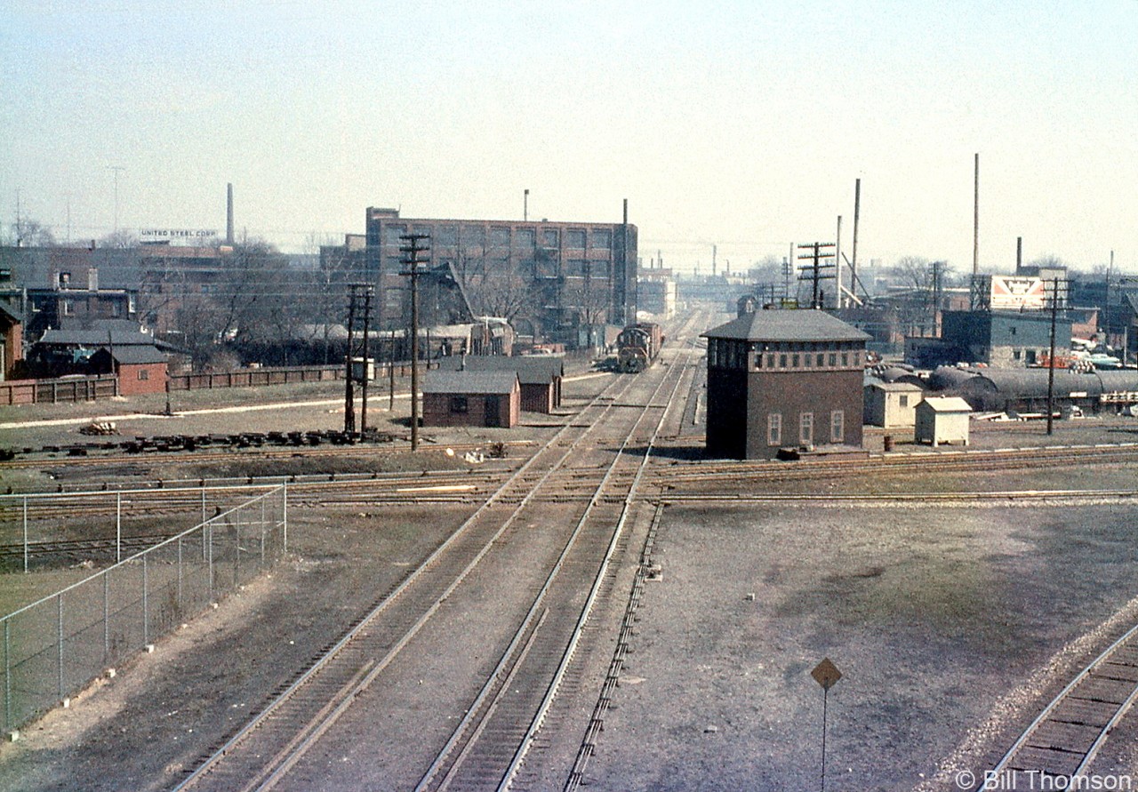 The CN-CP West Toronto diamonds in "The Junction" are pictured in 1963, looking east from the nearby Weston Road overpass. A short local lead by a CP Alco/MLW switcher is visible on the north mainline of the North Toronto Sub, near the switch to the MacTier Sub that headed north out of town for northern Ontario and the west coast. The West Toronto interlocking tower is prominently pictured on the south-east quadrant, and controlled movements over the diamond via a manual interlocking system (note the rods and linkages running from the base of the tower and along the tracks to control wayside signals and switches. The tower would be demolished next year in September 1964, and Centralized Traffic Control (CTC) installed and active by March 1965. A number of section houses dot the northeast quadrant opposite the tower.West Toronto was a main junction point for CP's lines leading north, east, and west out of the city of Toronto, as well as the location of their West Toronto & Lambton yards and locomotive servicing facilities. The closest track crossing CP's North Toronto Sub is CP's MacTier-Galt Sub connecting track, used regularly by the "The Canadian" between Toronto and Sudbury (via the MacTier Sub that lead north out of town) until the passenger train was rerouted by VIA the late 70's.The CP's Galt Sub is visible on the lower right, coming up from downtown Toronto (Union Station, and CP Parkdale Yard) and curving west to lead out of the city for Streetsville, Guelph Junction and lines beyond. The two main centre tracks running alongside the tower and crossing the diamond are for CN's Brampton Sub, which was their mainline from Toronto to Stratford until it was reorganized in January 1965. The Toronto-Halwest (near Bramalea) section became the Weston Sub, and after many decades this set of diamonds was grade separated in 2014.The furthest track crossing at the diamonds on the other side of the tower is the lead from the CP MacTier Sub to the "Old Bruce" service track that ran down the east side of the corridor to Dundas Street, serving local industries.CP's North Toronto Sub disappeared in the distance and cut through Toronto to Leaside, the Belleville Sub, and Agincourt (home of CP's new Toronto Yard that was under construction, opening next year). The opening of Toronto Yard meant much freight traffic and transfers would use the line between the two yards, and also to access the MacTier Sub and Galt Sub leading north and west out of town.
